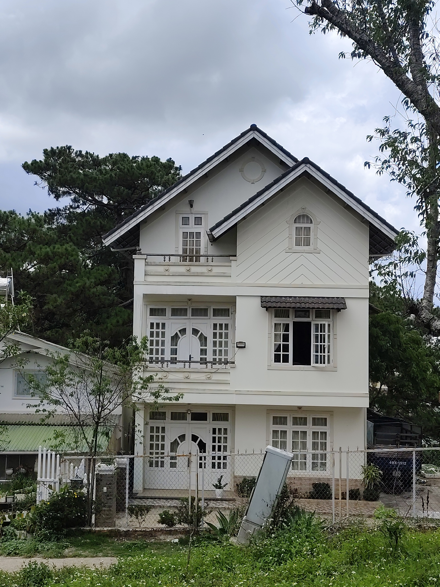 The image depicts a two-story white house with a dark roof and multiple windows behind a chain link fence in Da Lat, Vietnam. The house features architectural details such as a balcony, a semi-circular window, and angled siding. A smaller house is visible to the left. Trees and foliage surround the houses. The sky is overcast. An electrical box is angled and partially obscured by vegetation near the chain-link fence.