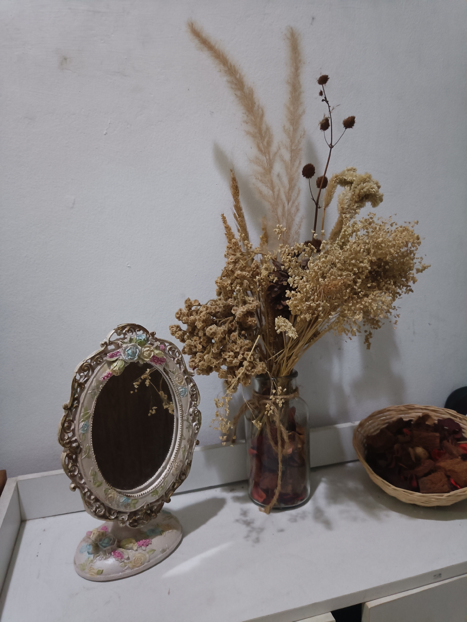 A still life arrangement is displayed on a white surface in West Jakarta, Indonesia. On the left, an oval mirror with an ornate, gilded frame decorated with floral motifs stands on a matching base. To the right of the mirror is a clear glass vase containing a bouquet of dried flowers and grasses, tied with twine. In front of the vase, within the vase, are dried red petals. To the right of the vase is a small, woven basket filled with dried potpourri. The background is a textured white wall. The lighting suggests an indoor setting with soft illumination creating shadows on the wall and surface.