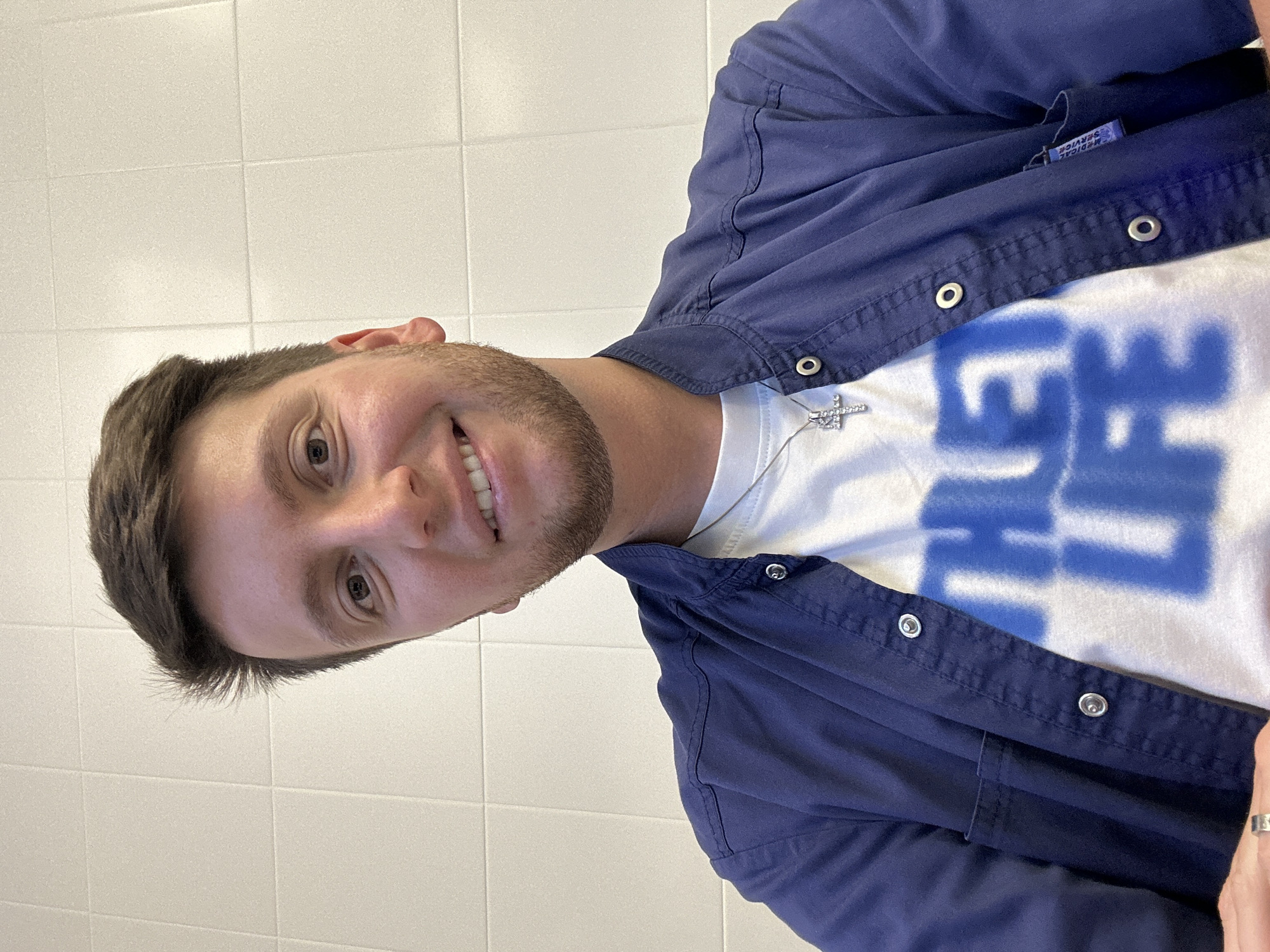A medium shot selfie taken from a slightly low angle shows a young man with fair skin, brown hair styled in a short, textured cut, and a light beard. He is wearing a white t-shirt with a large, blurry blue graphic that appears to read "THE LIFE", and an open, dark blue button-up shirt over it. A silver cross pendant necklace hangs from his neck. He is smiling directly at the camera. The background is a plain, cream-colored tiled wall. The man's hands are visible in the bottom left corner, with a silver ring on his right index finger. There is no indication of the specific location within Moscow, Russia.