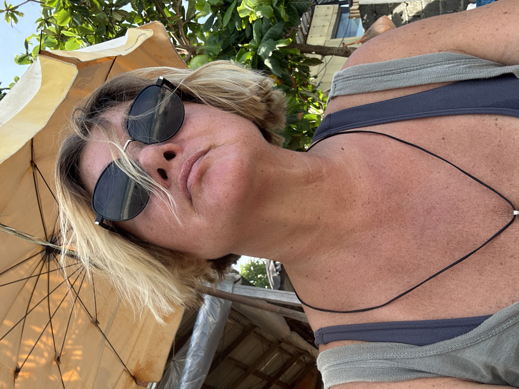 A close-up, low-angle selfie captures an adult with short, light blonde hair and dark sunglasses, looking downwards. The image is rotated, presenting a dynamic perspective of the subject's head and upper torso. The person's face is largely obscured by the round sunglasses, with only the nose, mouth, and chin visible, showing a relaxed or slightly pouting expression. Their skin appears tanned, with some redness on the neck and chest, suggesting sun exposure.
The person is wearing a light grey tank top or camisole over a dark blue undergarment, possibly a sports bra or bikini top. A thin black cord necklace with a small, light-colored pendant is visible on their chest.
To the left, a large, open beach umbrella or parasol in a light yellow or beige color dominates the foreground, providing shade. Its inner ribs and structure are clearly visible. Behind the subject, lush green foliage from trees or bushes forms a vibrant natural backdrop. In the background to the right, parts of a building or structure with a window and a corrugated metal roof are discernible.
The scene is brightly lit by natural sunlight, indicating a sunny daytime. The overall impression is one of leisure in an outdoor, possibly tropical, environment. The context indicates this location is Baan Chaweng, Thailand. There is no visible text in the image.