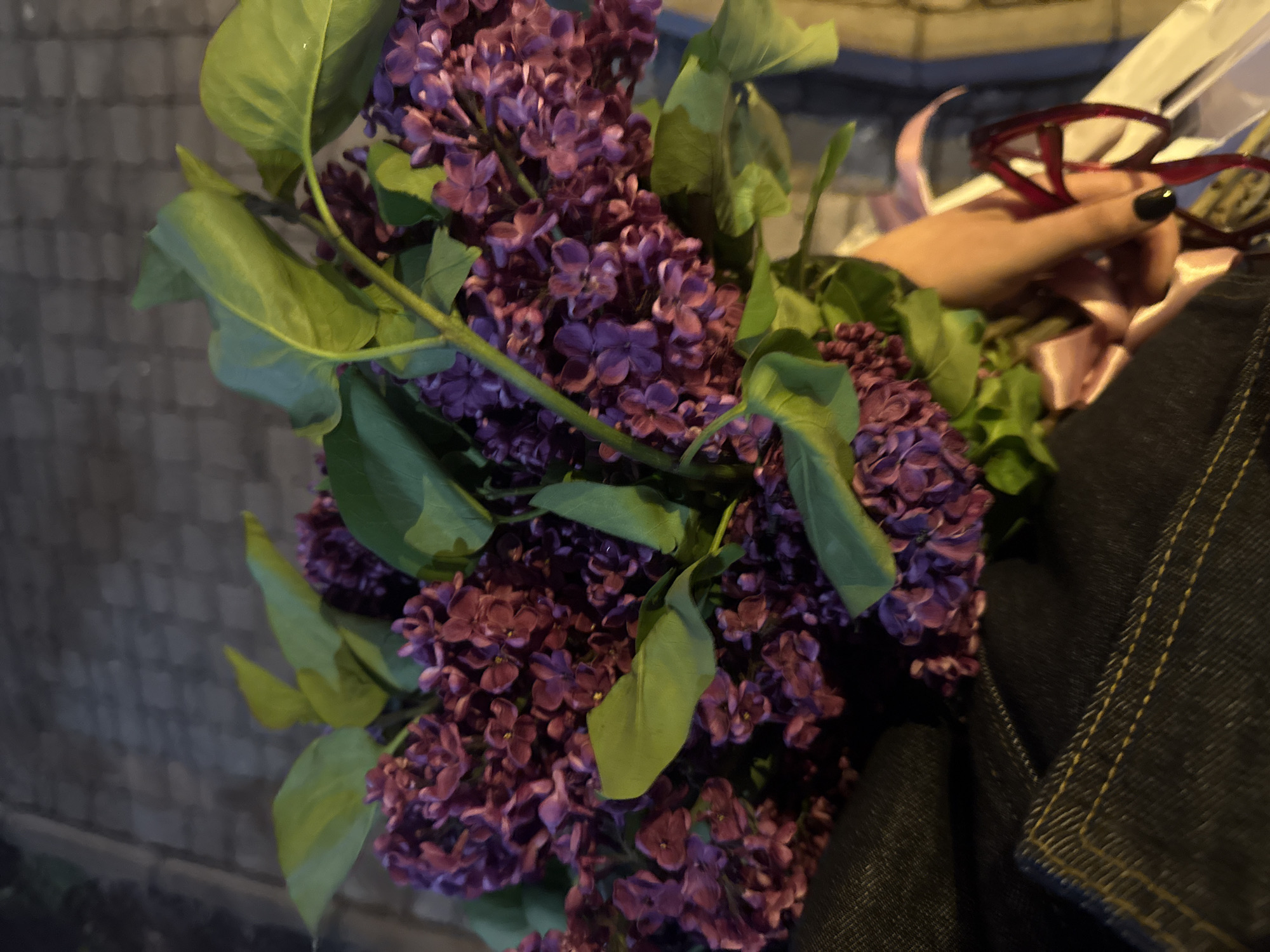 A close-up, slightly angled photograph depicts a bouquet of purple lilacs with green leaves. The lilacs are held in a hand with dark nail polish, also holding a pair of red-framed glasses wrapped with a pink ribbon. The background suggests an urban environment, potentially in Yerevan, Armenia, with an out-of-focus wall and pavement visible. The texture of a denim jacket can be seen at the edge of the photo.