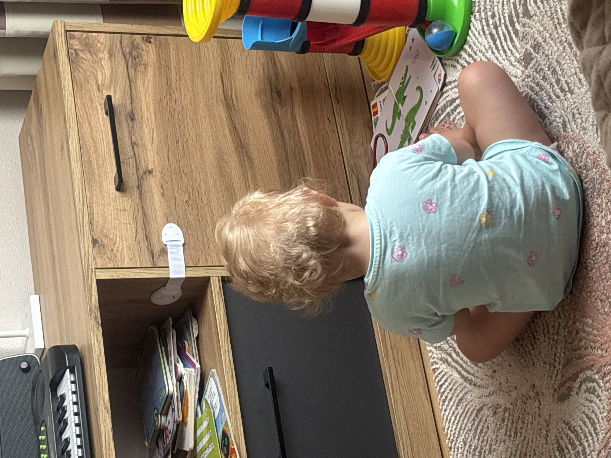 A young child with blonde, curly hair, viewed from behind, sits on a rug in front of a wooden cabinet. The child wears a light blue t-shirt with pink and yellow flower patterns. The cabinet has a light brown wood grain finish with a black handle on one drawer. A white safety latch is attached to the cabinet door. On the top shelf of the cabinet, to the left, is a black toy keyboard. To the right of the cabinet, a colorful toy structure with red, yellow, and blue components is visible. The floor is covered with a textured rug. A book with illustrations of green crocodiles is on the rug in front of the child. The scene appears to be indoors, possibly in a home environment. There is no discernible information about the specific location within Krāslava, Latvia.