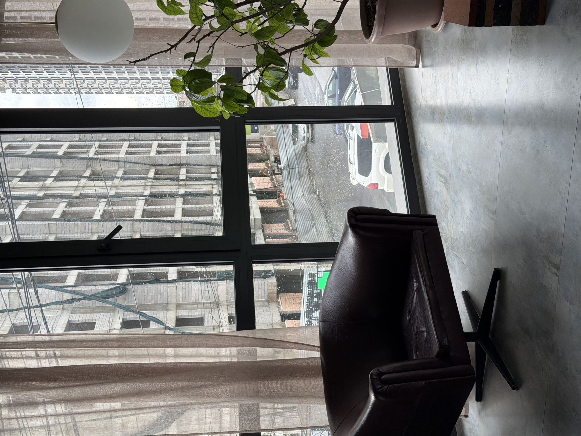 The image captures an indoor scene looking out through a large window onto an urban landscape, likely during daytime on an overcast or rainy day, given the wet windowpanes and street.
In the foreground, on the right, is a brown leather armchair with a distinctive black star-shaped base. The floor, visible in the lower portion of the image, is covered with light grey tiles. Sheer, light-brown curtains are partially drawn across the window, adding a soft, textured layer. A white, spherical light fixture hangs from the ceiling near the top left, and a potted plant with green leaves and thin branches is visible in the upper left corner, partially obscuring the view. Another plant pot is visible near the upper right edge.
Beyond the multi-paned window, an urban street scene unfolds. The window glass is dotted with raindrops or water streaks. Buildings, possibly under construction or with unfinished facades of concrete and bricks, dominate the view. Some structures show visible scaffolding or netting. On the street below, several cars are visible, including a white SUV or sedan and darker vehicles. The street appears wet, reflecting the light.
Visible text includes numbers "33" on a green sign and "22" on a small blue sign, both on distant buildings across the street. The overall impression is a quiet, contemplative view from an apartment or office in Yerevan, Armenia, looking out at a bustling, developing city.