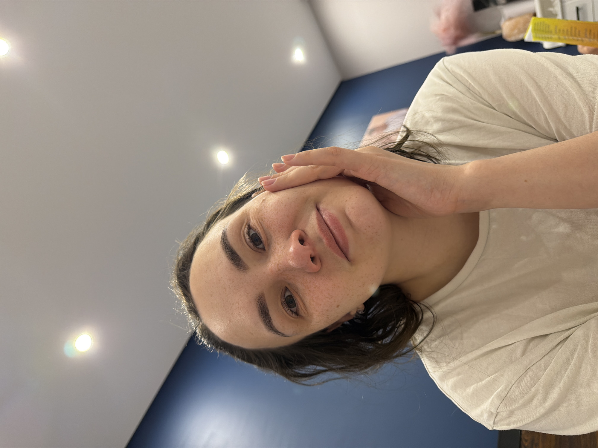 A woman with dark hair is holding her hand to her face, looking directly at the camera. She has fair skin with freckles. She is wearing a light-colored t-shirt. Behind her is a wall that is part blue and part white with several recessed lights. In the upper right corner of the frame, there are partially visible objects on a shelf. The location is Chaikovsky, Russia.