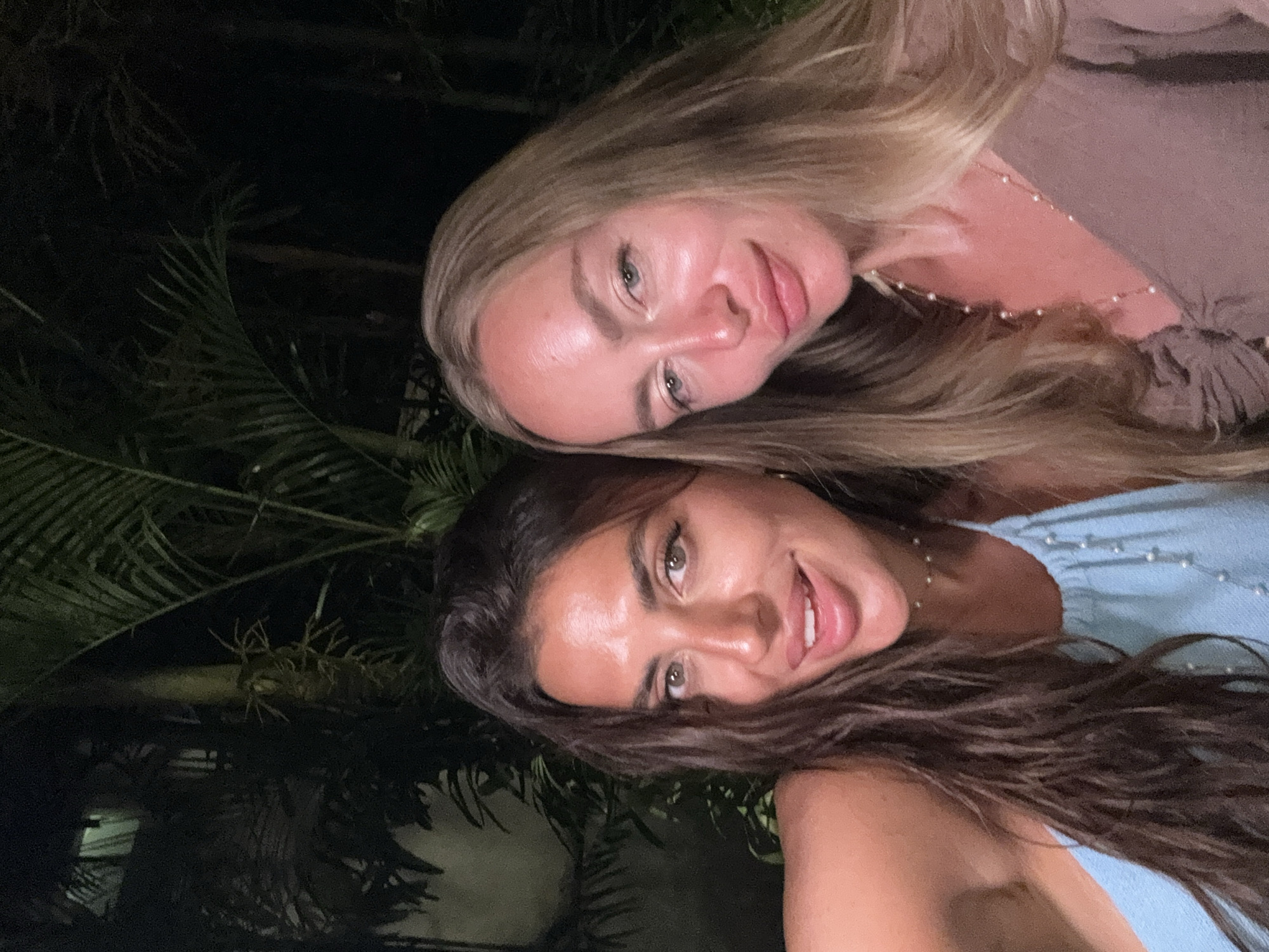 Two women are posing for a selfie-style photograph. The woman on top has long blonde hair and is wearing a light brown top and a delicate pearl necklace. The woman below has long, dark brown hair and is wearing a light blue top, also with pearl accents. Both women are smiling. The background is dark, with palm tree foliage visible. This photo appears to be taken outdoors at night. Given the location constraint of Kuta Selatan, Indonesia, the environment is likely a tropical or resort setting in the evening.