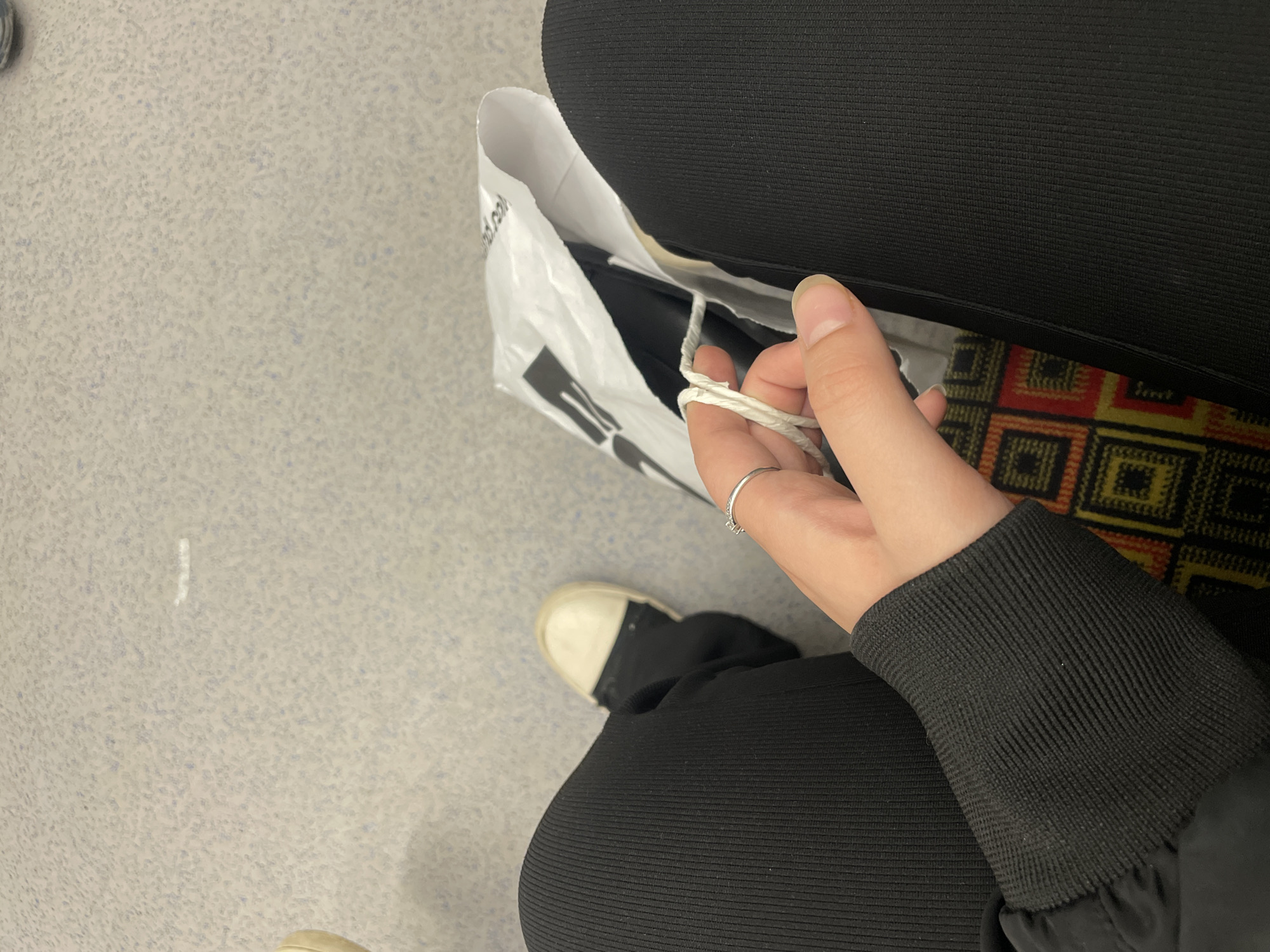 The image shows a high-angle, close-up view of a person indoors, focusing on their hand, legs, and a shopping bag. The person's left hand, adorned with a simple silver ring on the ring finger, is holding the twisted white rope handle of a crinkled white shopping bag. The person is dressed in black ribbed clothing, visible on their legs and as a cuff on their arm. A white and black shoe is partially visible towards the bottom left.
The setting features a light grey floor with small, speckled patterns, possibly linoleum or a similar commercial flooring material. To the right, a multi-colored patterned rug or carpet with geometric squares in shades of red, orange, yellow, and black is visible, suggesting an indoor public space.
The white shopping bag has faint, blurred black text and a logo on its surface; fragments like ".nd.co" can be partially discerned, but the full text is illegible. The scene captures a casual moment, possibly during or after a shopping activity, set in Saint Petersburg, Russia.