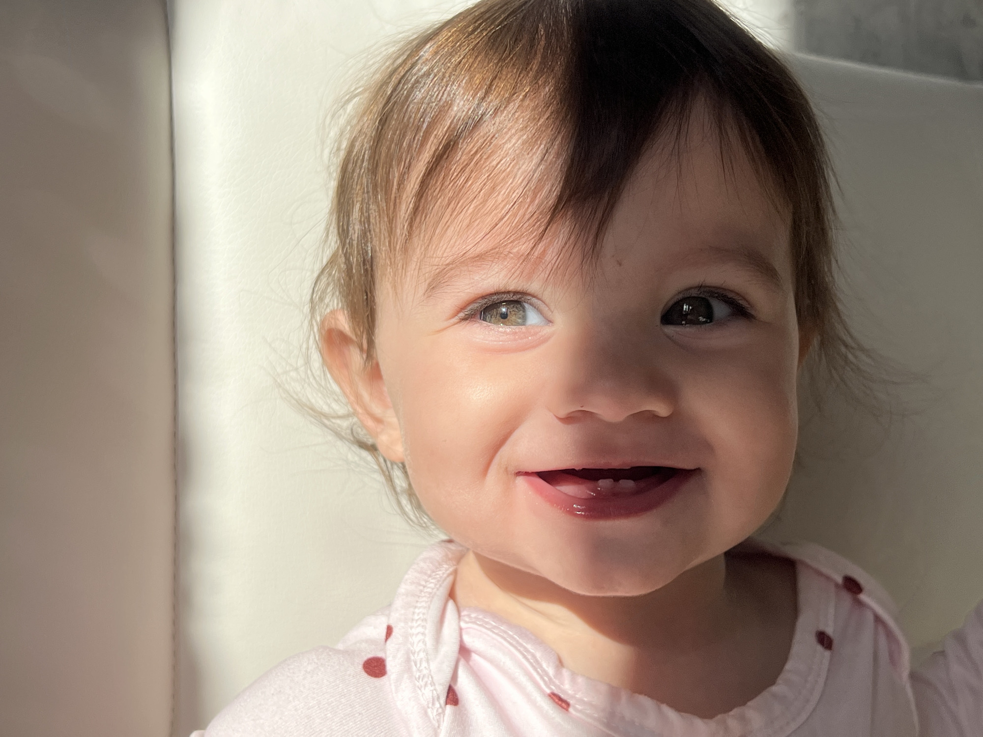 A close-up captures a smiling infant with light brown hair and hazel eyes. A shadow partially obscures the forehead and hair. The child wears a light pink shirt with small, darker pink polka dots. The background appears to be a white or light-colored surface, possibly a chair or wall. The image was not taken in Elena, Bulgaria as there is no information to suggest it was.