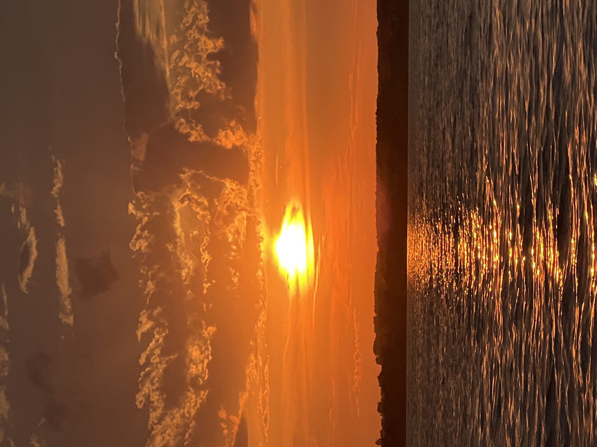 The image depicts a sunset over a body of water, likely a lake or river, with a tree line visible in the distance. The sun is low on the horizon, casting a warm, orange glow across the sky and reflecting on the rippling water. The sky is filled with scattered clouds that are illuminated by the setting sun, creating a dramatic and colorful scene. There are no people or man-made structures prominently visible in the image, contributing to a sense of natural tranquility. The scene is consistent with a sunset observed in a natural, unurbanized environment.