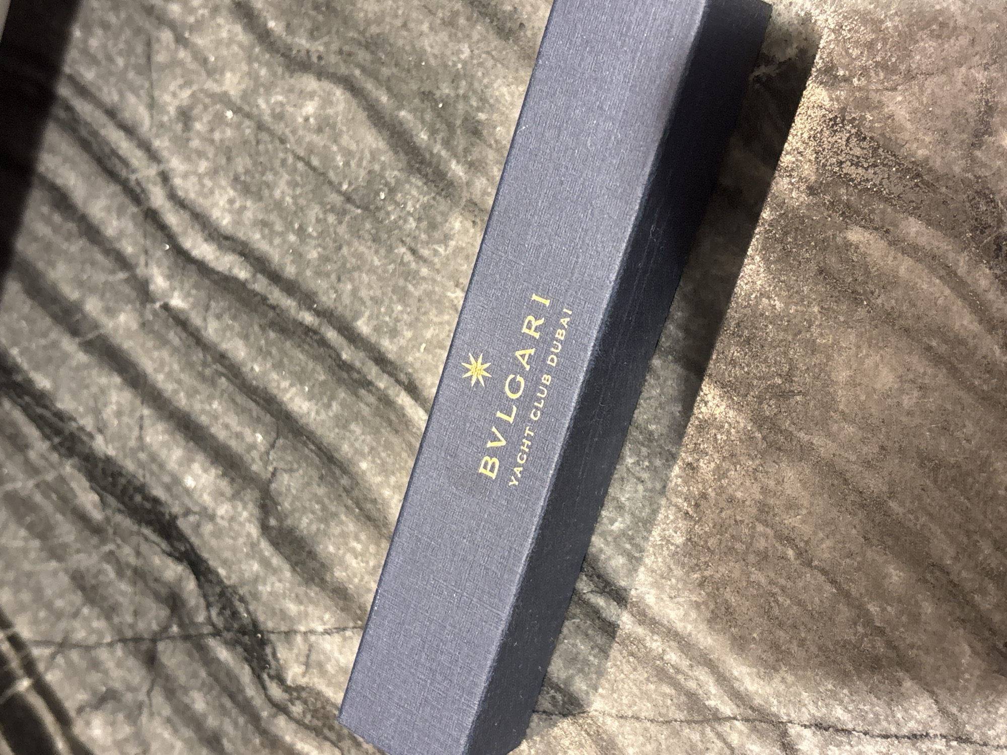 A navy blue rectangular box, likely a jewelry or gift box, rests on a speckled grey surface with dark grey veining. Gold lettering on the box reads "BVLGARI" with a star symbol above it, and below that, "YACHT CLUB DUBAI." The box is positioned at an angle, with its right side closer to the viewer. The background surface has a polished, marbled appearance, with irregular patterns of light and dark grey. The lighting suggests an indoor environment. There are no people or other objects visible in the image.