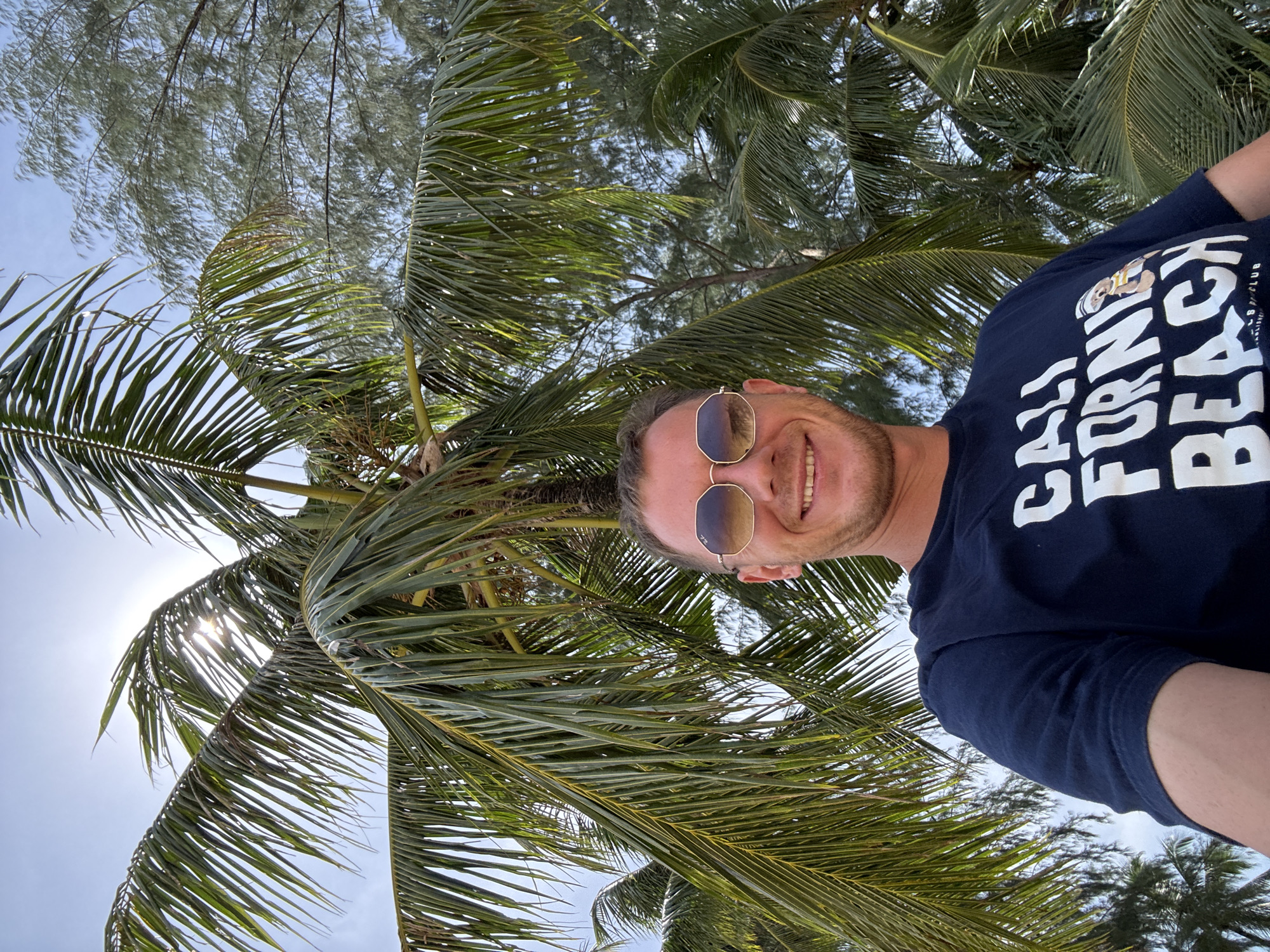 In Ban Khuk Khak, Thailand, a man with short brown hair and a beard is captured in a selfie. He is wearing sunglasses with reflective lenses and a dark blue t-shirt with white lettering that reads "CALI FORNIA BEACH". The man is smiling and positioned in front of lush green palm trees, with sunlight filtering through the fronds. The sky is visible in the background, appearing bright and clear. The setting suggests a tropical or coastal environment.