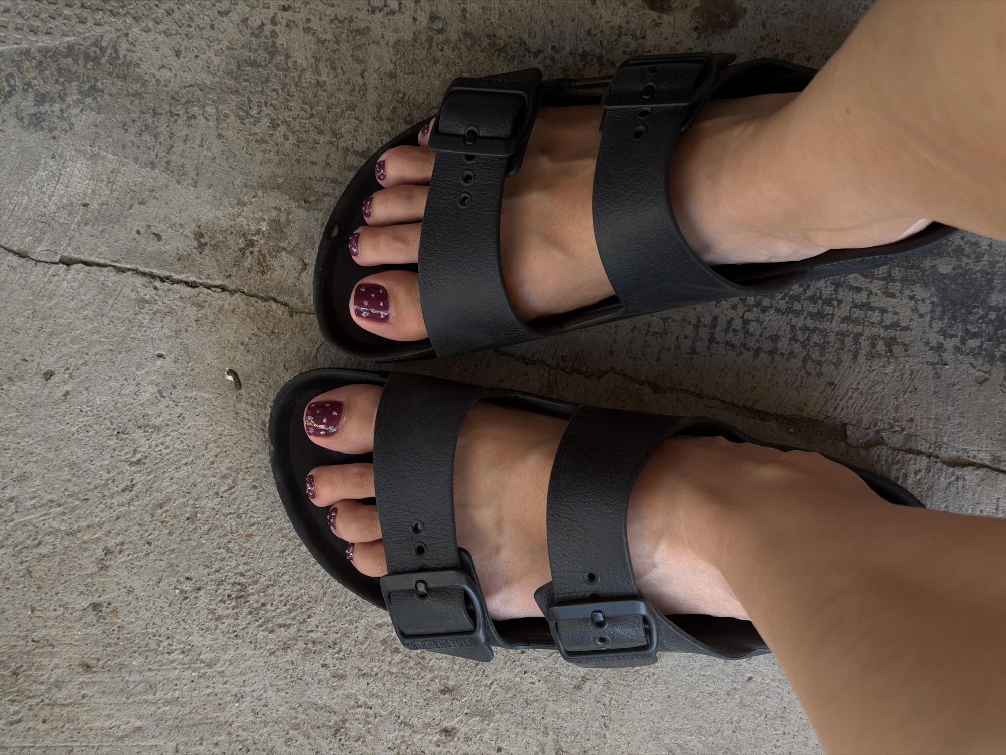 Viewed from above, the image presents a pair of feet clad in black Birkenstock Arizona sandals, resting on a textured concrete surface. The toes are visible, adorned with maroon-colored nail polish featuring a subtle glitter design. The sandals consist of two straps with adjustable buckles. The concrete ground appears to be cracked in some areas. The sandals are located in Nha Trang Ward, Vietnam.