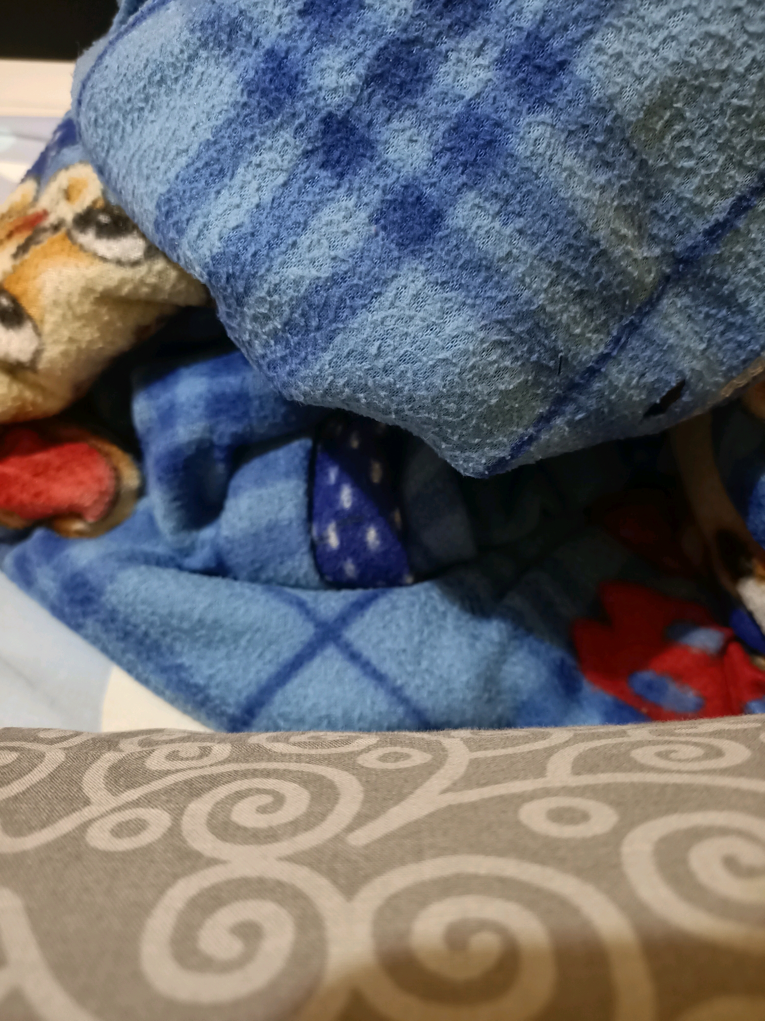 The image displays a close-up view of folded fabric. The most prominent element is a blue blanket with a textured surface, featuring a pattern of lighter blue and gray stripes. Portions of other fabrics are visible underneath and to the side. To the left, a glimpse of a light-colored fabric with a cartoonish animal face, possibly a fox, can be seen. Below this, a patch of red fabric with a black outline is visible. Further down, a section of a blue blanket with small white dots is present. In the foreground, a light-colored fabric with a repeating pattern of gray swirls occupies the bottom of the frame. The image is taken indoors, likely in a residential setting. There is no indication of the city of Perawang, Indonesia, in the image itself.