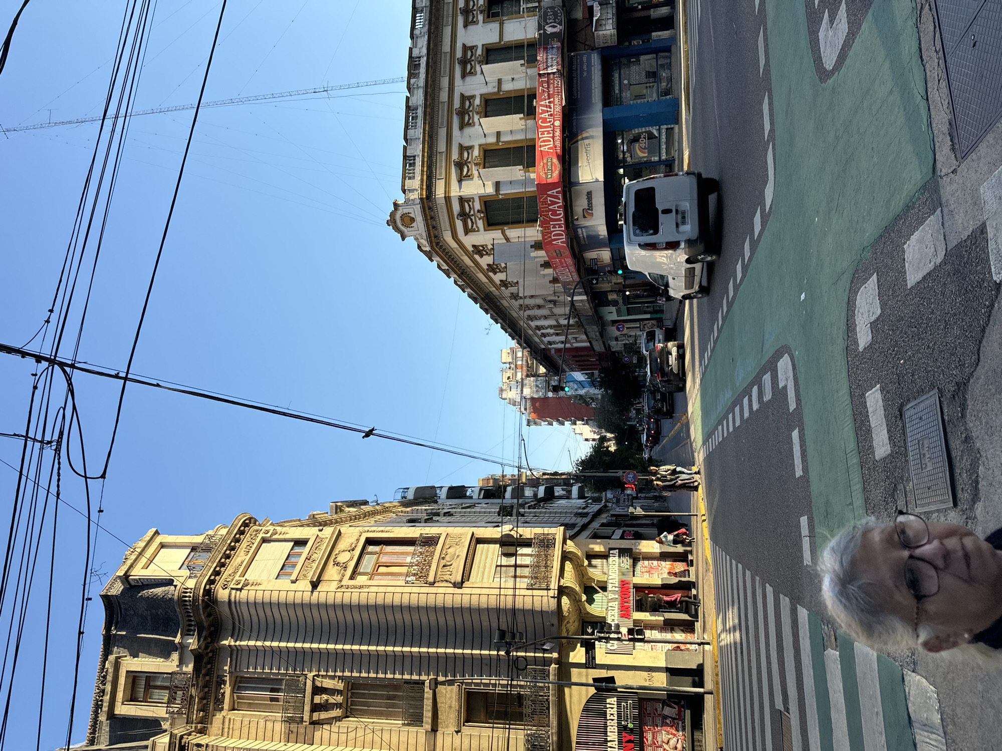 The image presents a daytime urban street scene in Buenos Aires, Argentina, characterized by its classical architecture and bustling activity. The perspective is looking down a city street lined with multi-story buildings featuring ornate facades and balconies. The sky above is clear and blue, crisscrossed by numerous overhead power lines and communication cables.
In the foreground, a green-painted bike lane is prominent on the right side of the road, marked with white dashed lines, running alongside a pedestrian zebra crossing. A white delivery van is visible in the car lane, with other vehicles and pedestrians further down the street. On the bottom right of the frame, the top of a person's head, with grey hair and wearing glasses, is partially visible, indicating a pedestrian's viewpoint.
The buildings display various commercial signs and banners. A large red banner on the right-hand building prominently features "ADELGAZA" (meaning "lose weight"), along with "WEI ZHEN" and several phone numbers: "11-3979-9641", "11-7368-0992", and "11-2254-39". Other visible text includes "Colchones Abaste" (likely a mattress supplier) with "Tel: 4863-0883", brand names like "Arspiris" and "Areotris", and a "40%" discount sign. A store on the left advertises "FIAMBRERIA" (delicatessen/cold cuts) and another sign likely reads "PANADERIA Y FIAMBRERIA" (bakery and delicatessen). The scene conveys the vibrant atmosphere of a typical sunny day in the city.