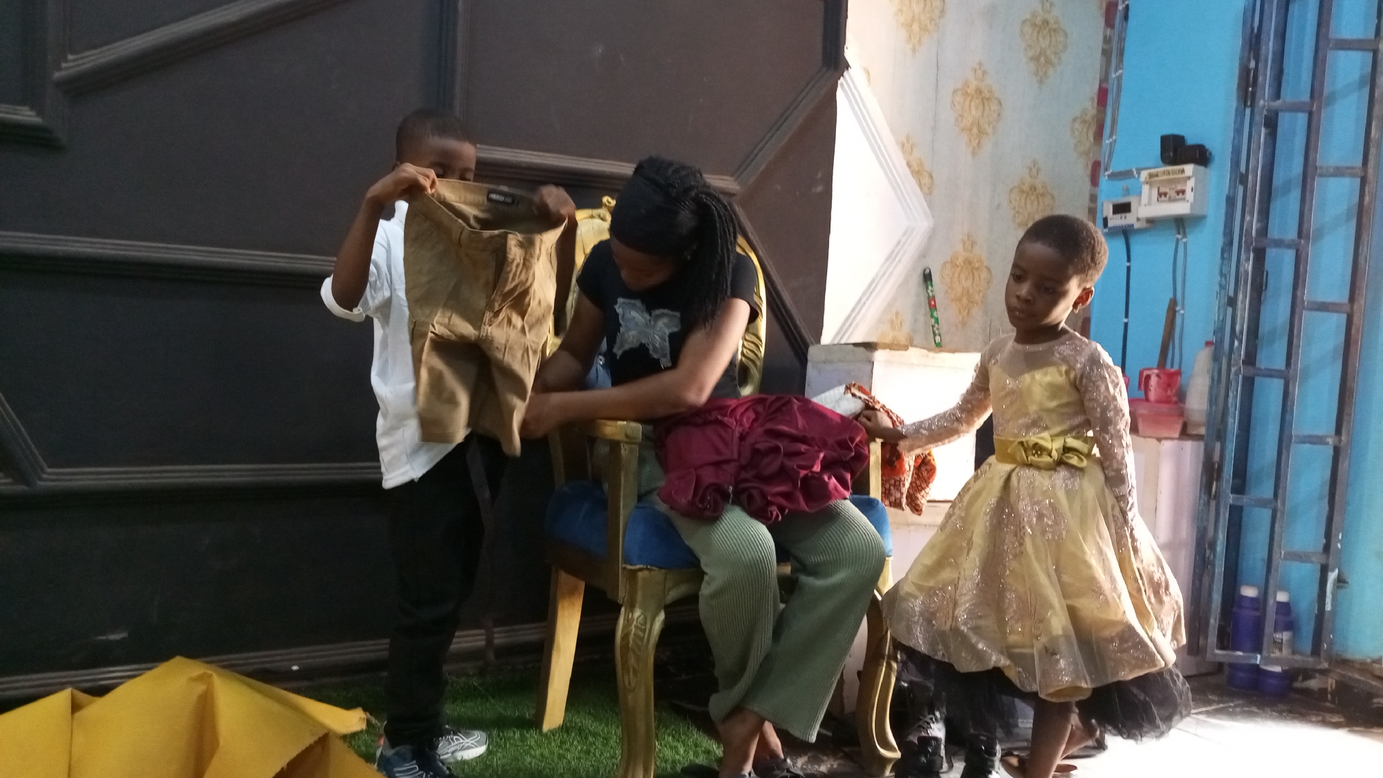 A woman sits in a gold-painted, blue-cushioned chair in what appears to be a staged or makeshift studio setting in Sabon Tasha, Nigeria. She wears a black t-shirt with a butterfly graphic, green pants, and a black head covering. She is looking down and appears to be handling a red, ruffled fabric. To her left stands a young boy in a white shirt and black pants holding up a pair of khaki-colored pants. To her right stands a young girl in a gold and black dress, holding the woman's hand. The backdrop consists of a dark, paneled wall on the left and a patterned wallpaper on the right. A metal window grate and electrical equipment are visible on the right side of the frame. A yellow fabric sits on the floor in the lower left corner. The floor appears to be covered with artificial grass.