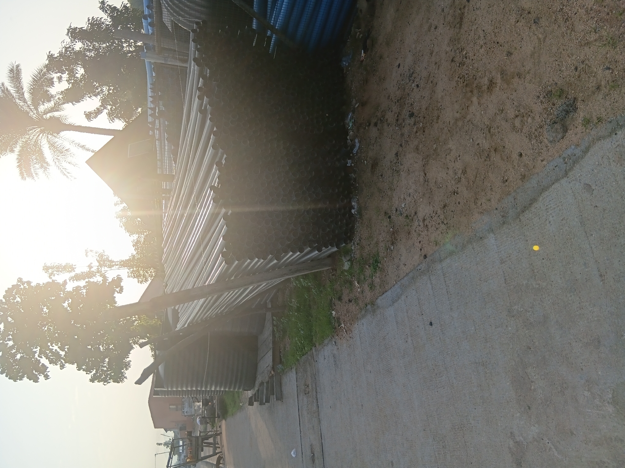 In Jalingo, Nigeria, a stockpile of black cylindrical pipes is displayed outdoors next to a paved path. The pipes are neatly stacked, with their circular openings clearly visible, forming a large, dark mass that dominates the right side of the image. Behind the pipes, a chain-link fence can be seen, and further back, a blue pipe is also visible. Sunlight beams down from the upper right, creating lens flare and casting a warm glow over the scene, highlighting a palm tree in the background. To the left of the pipes, a large, ribbed, dark-colored tank stands beside a wooden support structure. The ground is a mixture of sand and dirt. A concrete path runs along the left side of the image, with a few small green plants growing along its edge. In the distance, beyond a building, there is a hint of other structures and trees. The overall impression is an outdoor storage area for construction materials in a semi-arid environment.