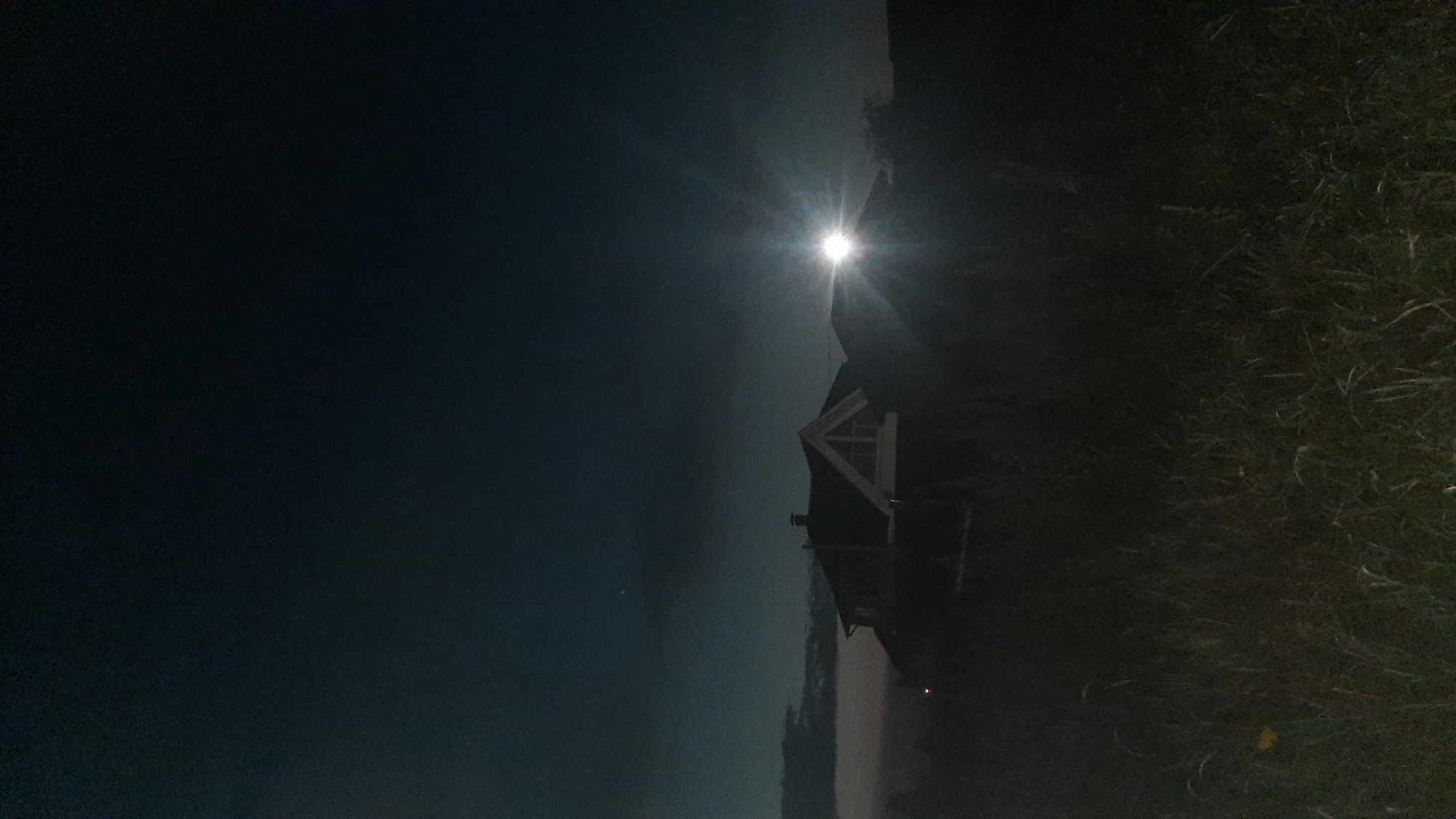 The image depicts a nighttime scene with a dimly lit house in the background. A bright light source, possibly a spotlight or a very bright lamp, is positioned to the right of the house, casting a strong glare that obscures details of the structure and its surroundings. The foreground is dominated by dark, tall grass, which appears somewhat overgrown. The sky is a deep, dark blue with some faint clouds visible. The overall impression is one of a rural or suburban setting at night, with artificial light creating a dramatic effect. There are no people visible in the image. The location in Shklov, Belarus, is not discernible from the visual information provided in the image.
