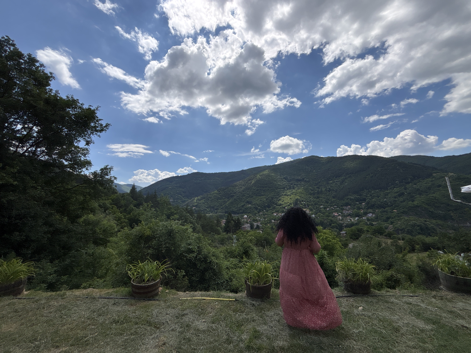 A woman with long, dark, curly hair stands with her back to the camera, wearing a long, flowing, pink dress with a subtle pattern. She is looking out over a vast, verdant valley filled with rolling hills covered in lush green trees. In the distance, a small village is nestled amongst the trees, with buildings visible amongst the foliage. The sky above is a vibrant blue, dotted with dramatic, fluffy white clouds that cast shadows on the landscape.
The setting appears to be an elevated viewpoint, possibly a terrace or balcony, with a grassy foreground. Two large potted plants, filled with spiky green foliage, flank the woman. A yellow hose or cable lies on the grass between the pots. The overall atmosphere is peaceful and serene, with a sense of awe inspired by the expansive natural scenery. The time of day appears to be daytime, with bright sunlight illuminating the scene and creating a contrast between light and shadow. The weather seems to be fair, with a mix of sun and clouds. The location is identified as Lukovo, Bulgaria. No text is visible in the image.