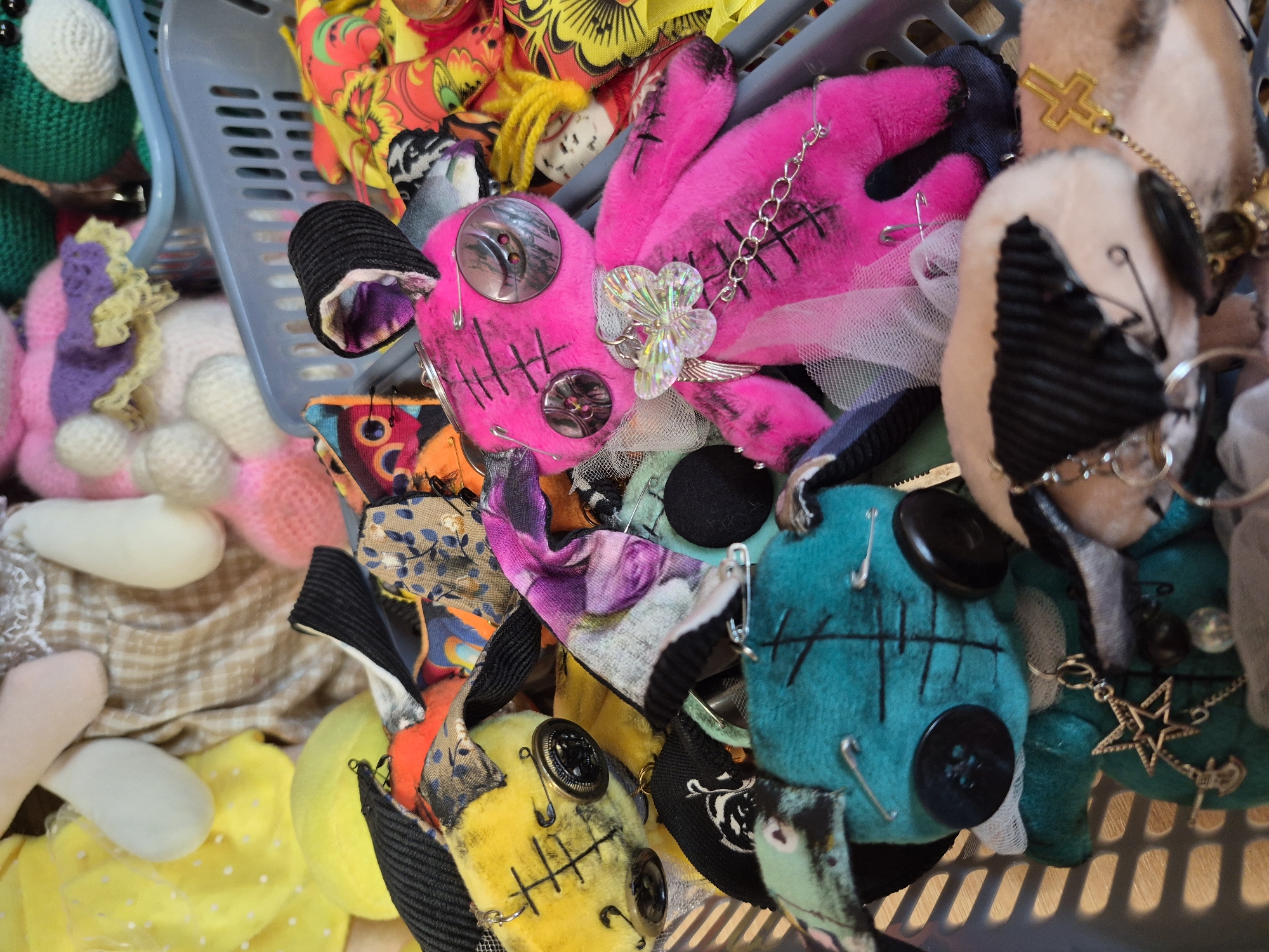The image depicts a collection of handmade plush toys, commonly referred to as "creepy dolls," arranged within a light gray plastic basket. The dolls are characterized by their somewhat unsettling aesthetic, featuring mismatched button eyes, stitched facial features, and exposed seams. One prominent doll is bright pink, adorned with large, reflective button eyes, stitch marks, and a delicate butterfly charm hanging from a chain around its neck. Another doll, teal in color, also has button eyes and a star-shaped charm dangling from its neck. Other dolls visible in the image are yellow and feature similar construction. The dolls are interspersed with various fabric scraps and embellishments, including safety pins, ribbons, and translucent mesh. The background suggests a cluttered environment, with other plush toys and fabric items partially visible. The lighting is moderate, casting soft shadows on the subjects. The overall scene appears to be an indoor setting, possibly a craft area or a display of handmade items. There is no discernible information in the image that can indicate its location in the city of Красный Ключ, Russia.