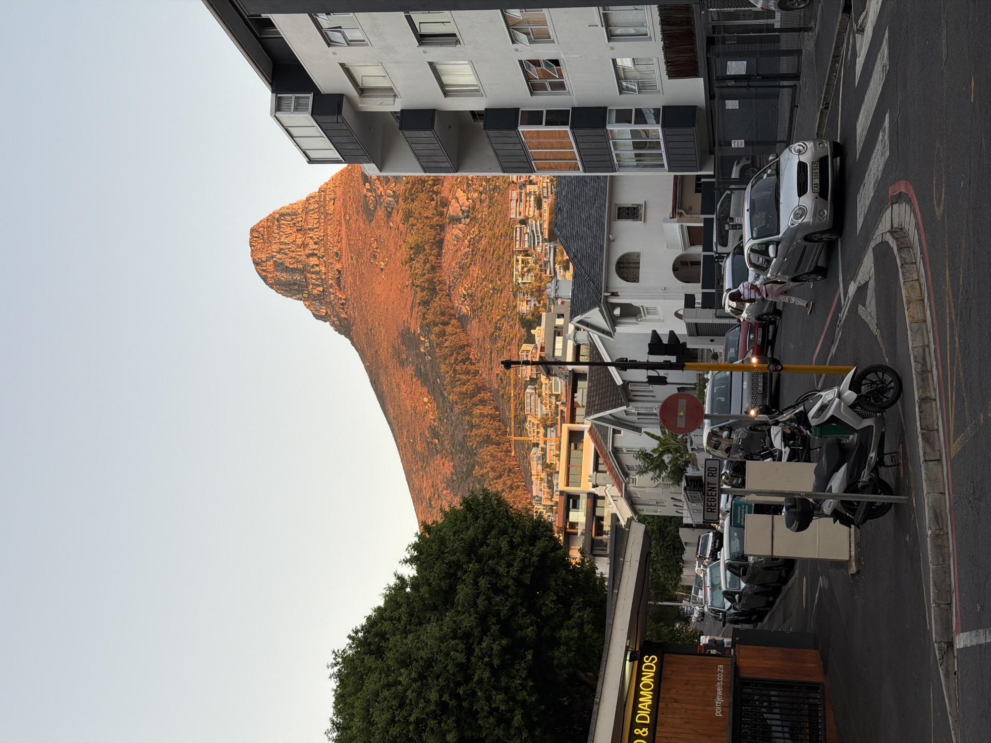 The image shows a street scene in Cape Town, South Africa, specifically Regent Road. Prominently displayed in the background is Lion's Head mountain.
Buildings line the street, varying in height and architectural style. A multistory building with white walls and black balconies is on the left side of the frame.
Several vehicles, including cars and motorcycles, are parked along the side of the road. One person is seen standing near a silver car.
Street signs are visible, including one reading "Regent Rd". A shop sign saying "& Diamonds" is also visible.
The sky is pale blue, suggesting either dawn or dusk, with the mountain illuminated with a warm, orange glow.