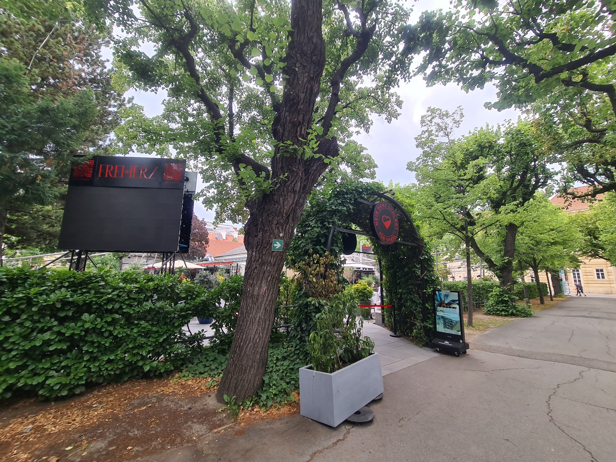 The image shows an outdoor scene in Vienna, Austria, likely at the Prater amusement park. On the left, a large black digital billboard displays the word "FREIHERZ" in red lettering with a textured background. Behind the billboard is a neatly trimmed green hedge. To the right of the billboard, a large tree trunk is prominent, with lush green foliage from the trees above.
Further to the right, an arched entryway is covered in dense green ivy, forming a tunnel-like structure. Inside the arch, a round sign with a red heart and the text "EIN" and "FÜR STIEGL" is visible. To the right of the ivy-covered arch, a tall, black digital display screen on wheels shows an advertisement. The ground is paved with concrete, and there are fallen leaves on the dirt path to the left of the tree. In the background, more trees, buildings with reddish-tiled roofs, and a street can be seen, with a few people walking in the distance. The sky is overcast.