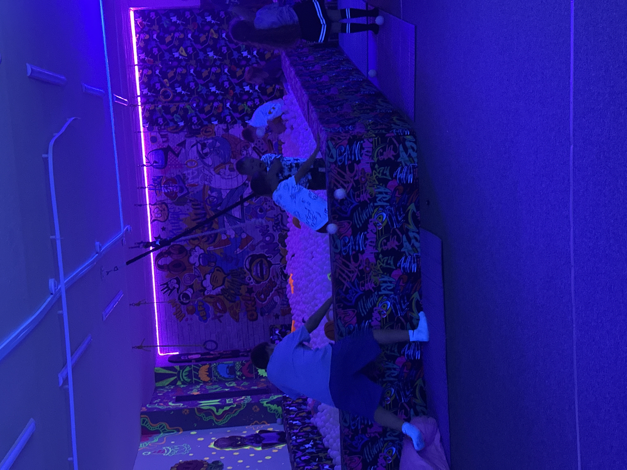 The image shows an indoor recreational space, possibly a children's play area, illuminated with ultraviolet or black lights. The walls are decorated with colorful, graffiti-style artwork. Several people, including children and an adult, are present and appear to be playing in a large pit filled with white balls. Some are standing on the edge of the pit. The floor and various surfaces are covered in patterned, fluorescent designs that glow under the UV light. The exact city location is unknown.
