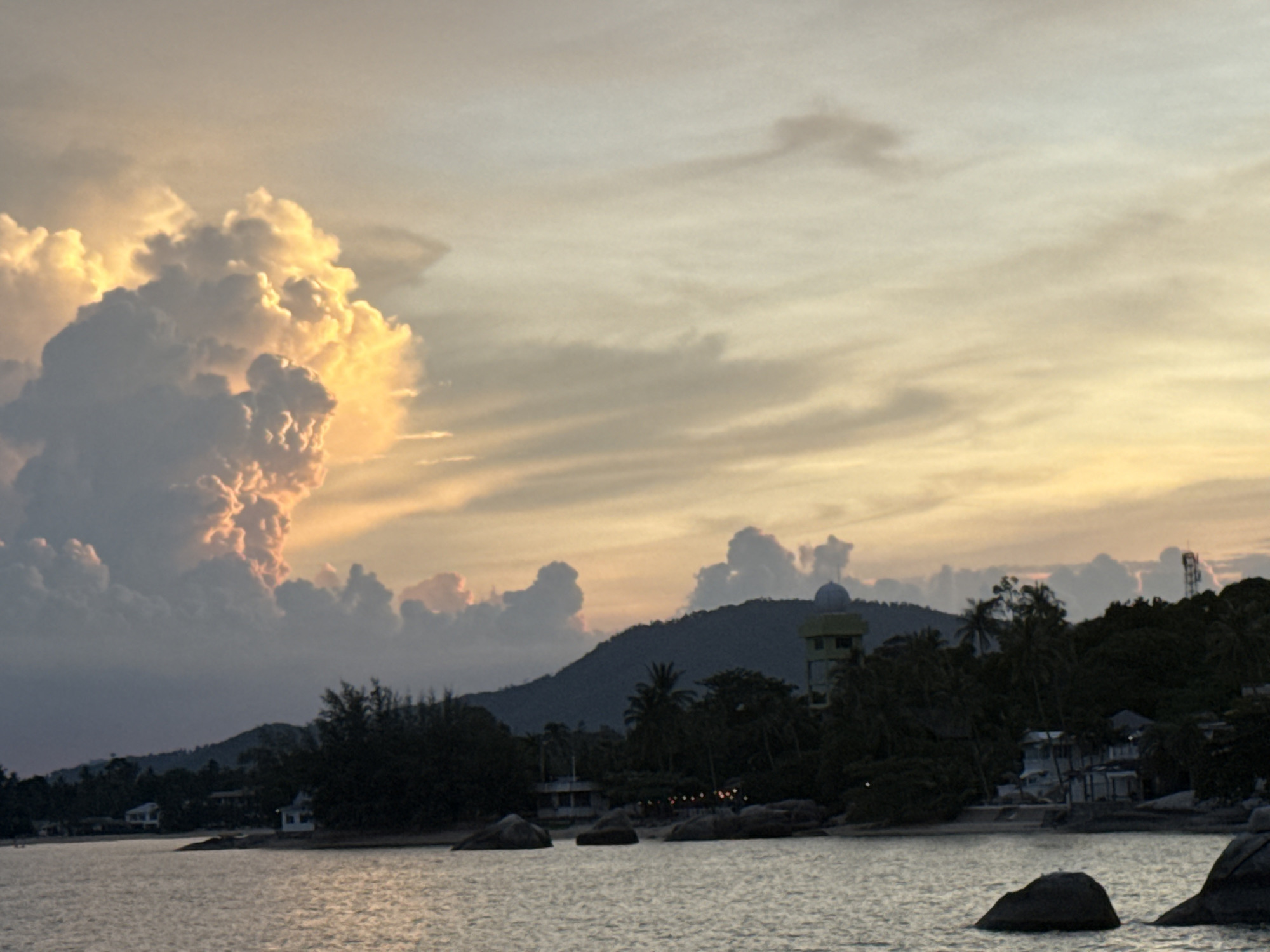 The image captures a coastal scene at sunset in Baan Hua Thanon, Thailand. The sky is filled with dramatic clouds, illuminated by the setting sun, with hues of orange, yellow, and soft white. In the foreground, the calm ocean surface reflects the colors of the sky. The shoreline is dotted with large rocks and boulders. Behind the rocks, a stretch of land is visible, characterized by lush green trees and vegetation. Several buildings, appearing to be houses or small structures, are nestled among the trees. A prominent feature on a hilltop is a green tower with a dome-like structure. The overall atmosphere is serene and natural, typical of a coastal village setting.