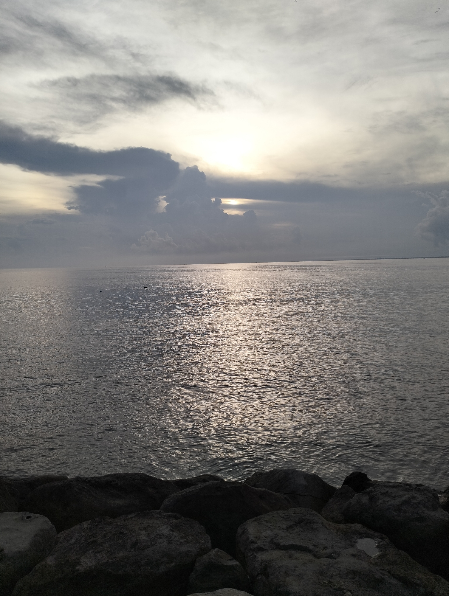 The image depicts a seascape at Lhokseumawe, Indonesia. The sky is overcast with gray clouds, partially obscuring the sun. The sea surface reflects the light, creating a shimmering effect. In the foreground, large dark rocks are visible along the shoreline. The overall scene suggests a tranquil, potentially overcast day at the coast.