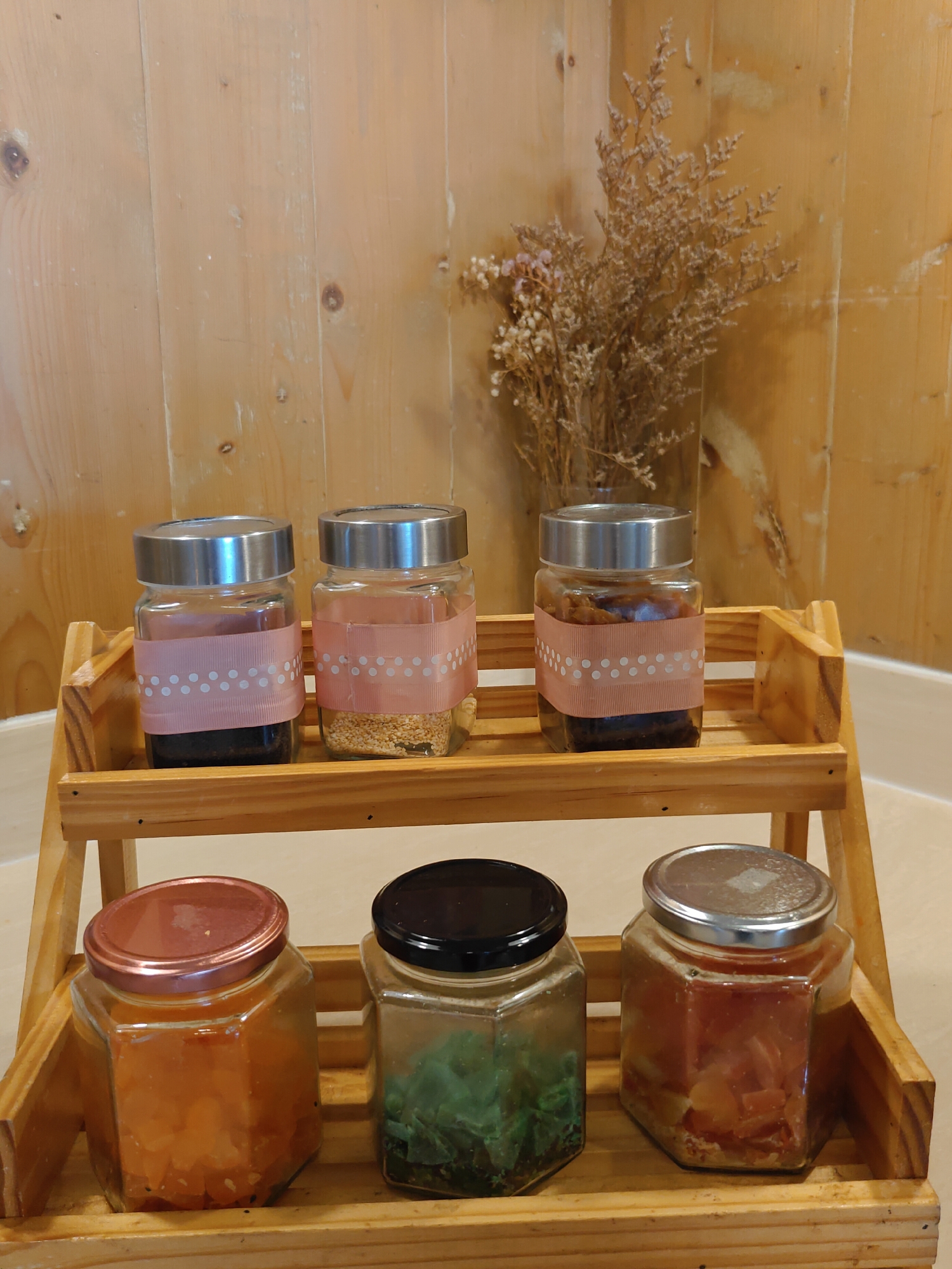 The image depicts a two-tiered wooden shelf unit positioned against a light brown wooden plank wall. On the top shelf, three glass jars with metallic lids are arranged. Each jar is adorned with a pink ribbon featuring a white polka dot pattern. The contents of these jars appear to be liquids and grains. Behind the jars on the top shelf, a vase containing dried floral arrangements is visible against the wooden wall.
The bottom shelf holds three hexagonal glass jars. The leftmost jar has a rose-gold colored lid and contains an orange-colored substance. The middle jar has a black lid and contains green leafy material. The rightmost jar has a metallic lid and is filled with pieces of fruit. The overall setting suggests a kitchen or pantry area. There are no people or animals present in the image. The image was taken in Pattaya City, Thailand.