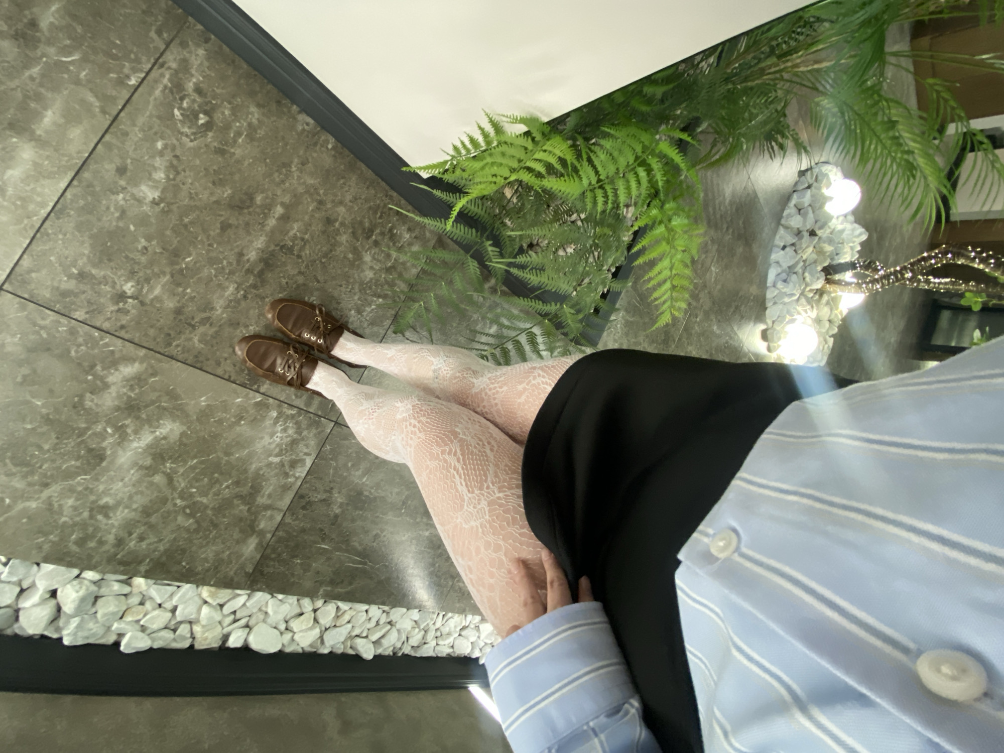 A first-person perspective photograph shows a person's legs clad in patterned white tights, wearing brown lace-up shoes, a black skirt, and a light blue, white-striped button-down shirt. The background consists of gray marble flooring and a border of small white stones along the bottom left. A potted fern is positioned adjacent to a decorative display of white stones and string lights. The location is not discernable within the city of Votkinsk, Russia based on the visual elements present.