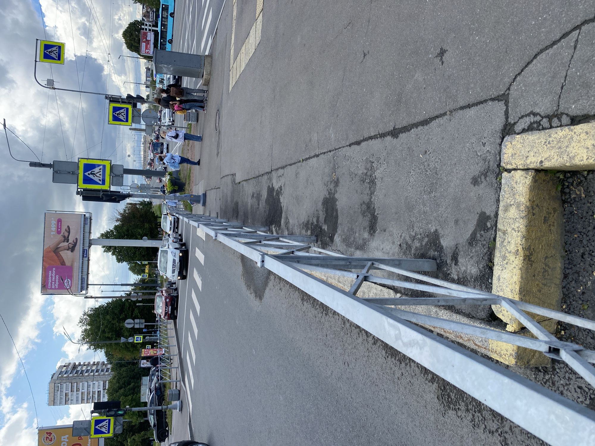 The image shows a street scene in Saint Petersburg, Russia, likely near a crosswalk. In the foreground, a metal fence runs along the side of the road, bordering a sidewalk. The sidewalk is made of asphalt and shows some wear and tear, with visible cracks and dark patches.
Several pedestrian crossing signs, featuring a blue square with a white pedestrian silhouette, are visible, indicating a designated crosswalk. Traffic lights are also present. On the sidewalk, a group of people are waiting, some facing the street, suggesting they are waiting to cross or have just crossed.
The street is paved with asphalt and marked with white stripes for the crosswalk. Cars are present in the background, with one red SUV and a white sedan stopped at the crosswalk. A blue bus is visible further down the road.
Several tall buildings are visible in the background, characteristic of urban architecture in Saint Petersburg. Large billboards are also present, one of which appears to advertise sandals. The sky is partly cloudy. The scene suggests an active urban environment with pedestrian and vehicular traffic.