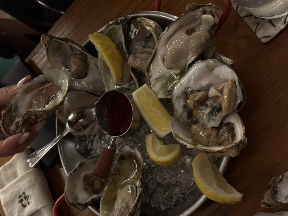 A close-up, high-angle view depicts a circular metal platter of shucked oysters on crushed ice, resting on a dark reddish-brown wooden table in a dining establishment in Canggu, Indonesia. Approximately ten oysters are visible in their half-shells, some appearing plump and glistening with natural liquor. Several yellow lemon wedges are distributed among the oysters. A small, stainless steel saucepot with a dark wooden handle contains a dark red sauce, positioned centrally on the ice. A silver spoon with an ornate handle lies on the ice near the saucepot. In the upper center, a person's hand, exhibiting manicured fingernails, holds one of the oysters by its shell. A light-colored fabric item with a dark embroidered symbol is partially visible in the upper left background, and a white and green patterned coaster is partially visible in the lower right. The ambient lighting is dim.