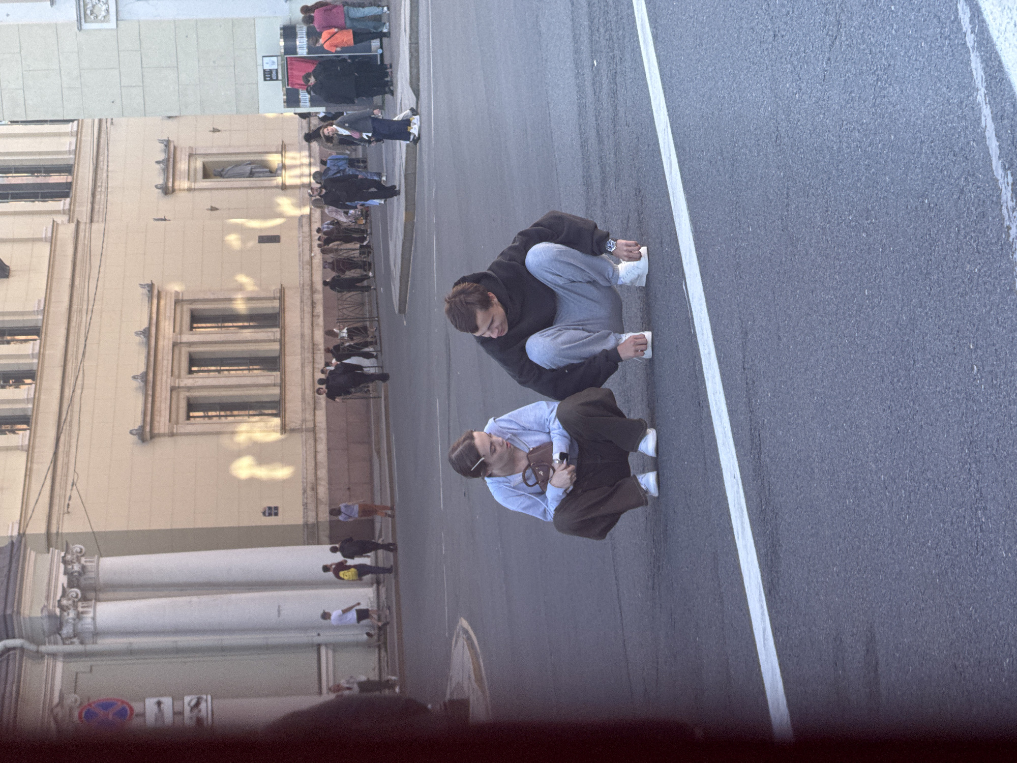 The image depicts a street scene in Saint Petersburg, Russia. Two young individuals are crouched down in the middle of an asphalt road, facing each other. The person on the left, a female with light brown hair pulled back and a hair clip, wears a light blue hooded sweatshirt and dark brown wide-leg trousers. She holds a small brown handbag. The person on the right, a male with short brown hair, wears a dark grey hooded sweatshirt and light grey sweatpants. Both are wearing white sneakers.
In the background, a group of people are walking on the sidewalk. Beyond the sidewalk, a light-colored building with architectural details, including columns and windows, is visible. The building appears to be an older structure, characteristic of city architecture. Sunlight casts shadows on the building's facade. Traffic signs are present on the left side of the image. The overall atmosphere suggests a public space in the city during daylight hours, possibly late afternoon given the lighting.