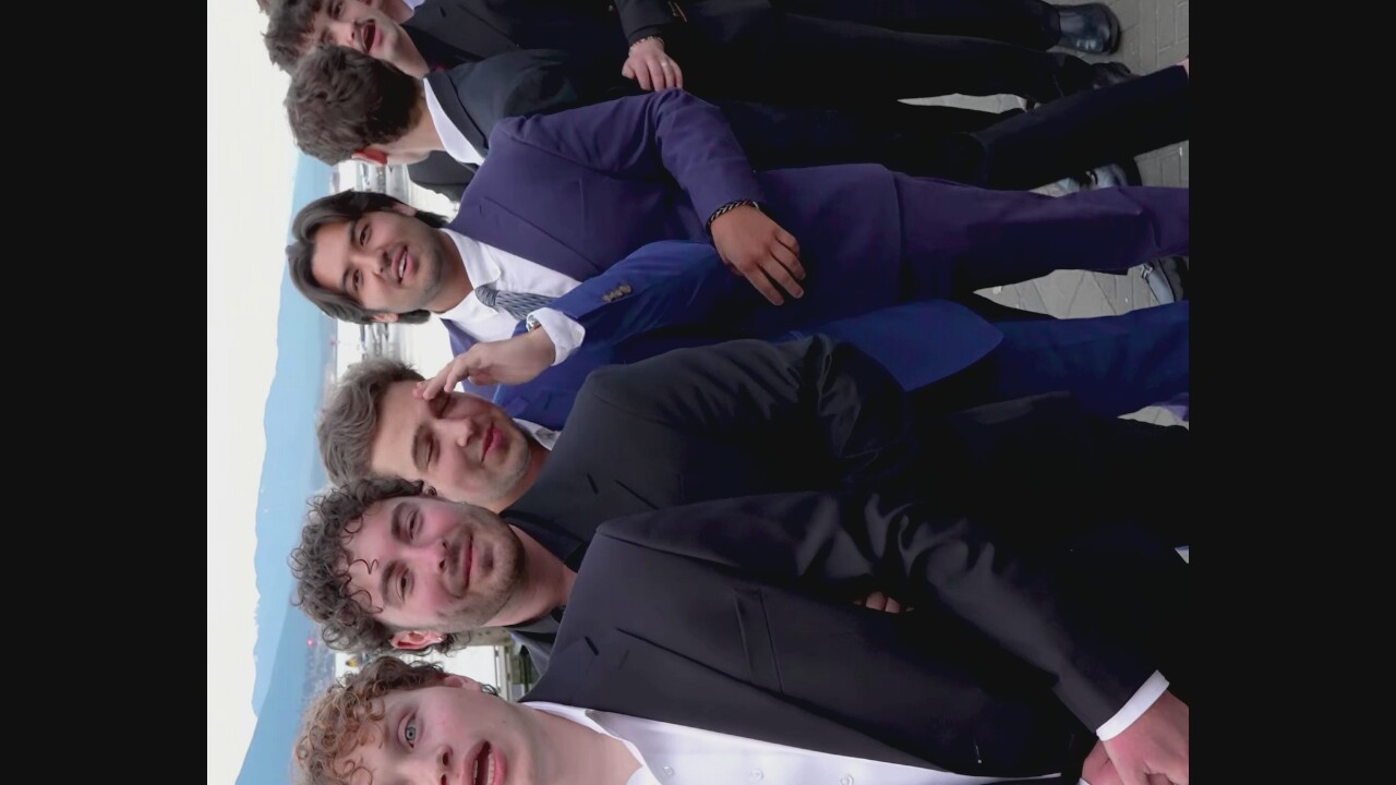 Six young men in formal attire are posing for a photograph outdoors. The men are standing in a line, facing the camera. The man on the far left is wearing a black suit, a white dress shirt, and is looking towards the camera with his mouth open. The next man to his right is wearing a black suit with a dark shirt and is smiling. The third man from the left is wearing a dark suit and appears to be touching his forehead. The fourth man is wearing a blue suit and tie, looking towards the camera with a slight smile and his hand on his forehead. The fifth man from the left is wearing a dark suit and is partially obscured. The sixth man from the left is also wearing a dark suit and is partially obscured.
The background suggests an outdoor setting with a body of water and mountains in the distance, consistent with a location in Vancouver, Canada. The ground beneath the men is paved, possibly a boardwalk or promenade. The lighting indicates it is daytime.