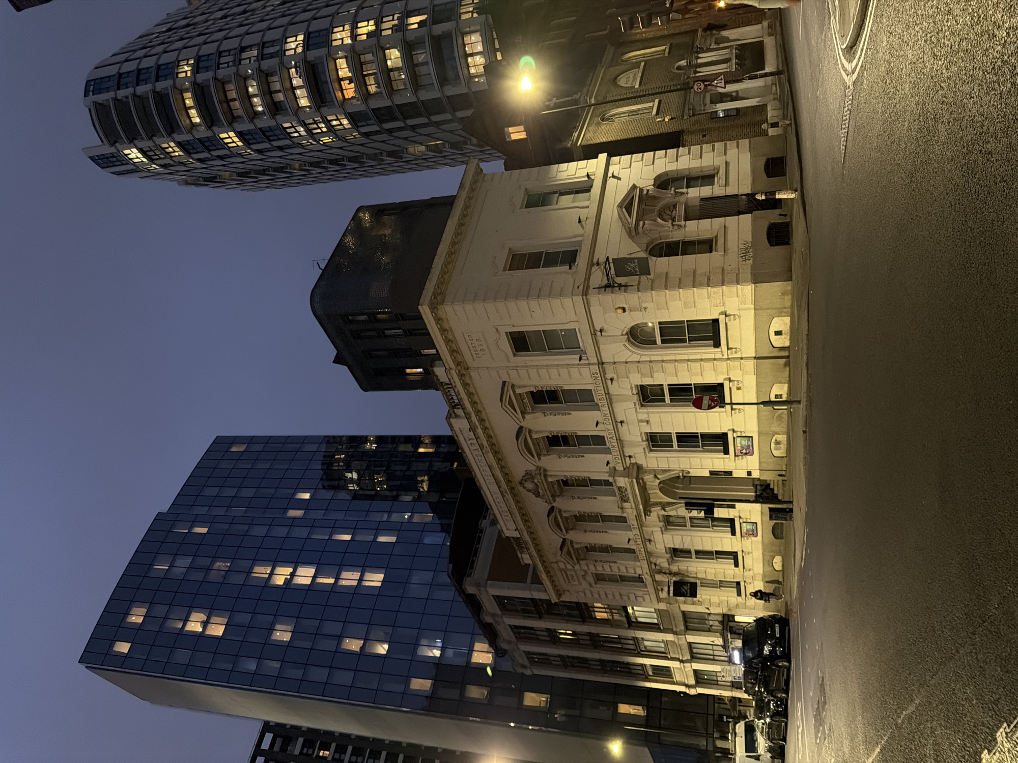 An eye-level view on a city street in London, United Kingdom, at dusk or night. The main subject is a multi-story white building with classical architectural details, including arched windows and decorative moldings. Adjacent to it is a modern glass skyscraper with numerous lighted windows, reflecting an urban environment. A taller building, curved and with multiple lighted windows, is also visible in the upper left. The street is illuminated by streetlights, which cast shadows and highlight the texture of the pavement. Parked cars are visible along the curb. There appear to be a few people walking near the building.