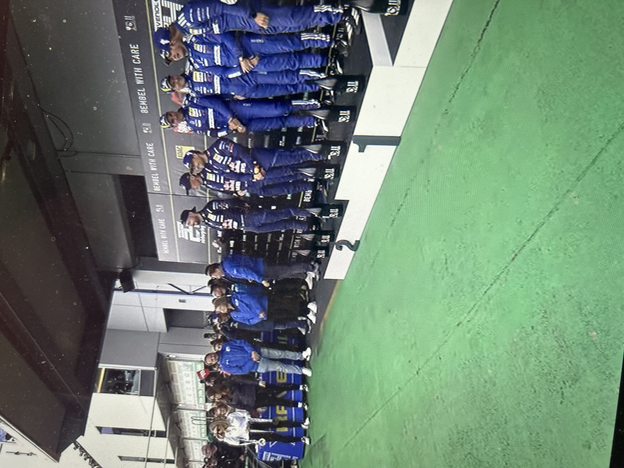 The image shows a large group of people posing on a tiered podium that has the numbers 1, 2, and 3 on the front. The people on the top tier are wearing blue racing suits and hats. The lower tiers include people in varying street clothes such as jackets, jeans, and hats. The backdrop behind the podium features text including "BEMBEL WITH CARE." The area appears to be part of an indoor or covered structure, possibly a garage or pit area. The location is unknown and can not be inferred to be in Sofia, Bulgaria.