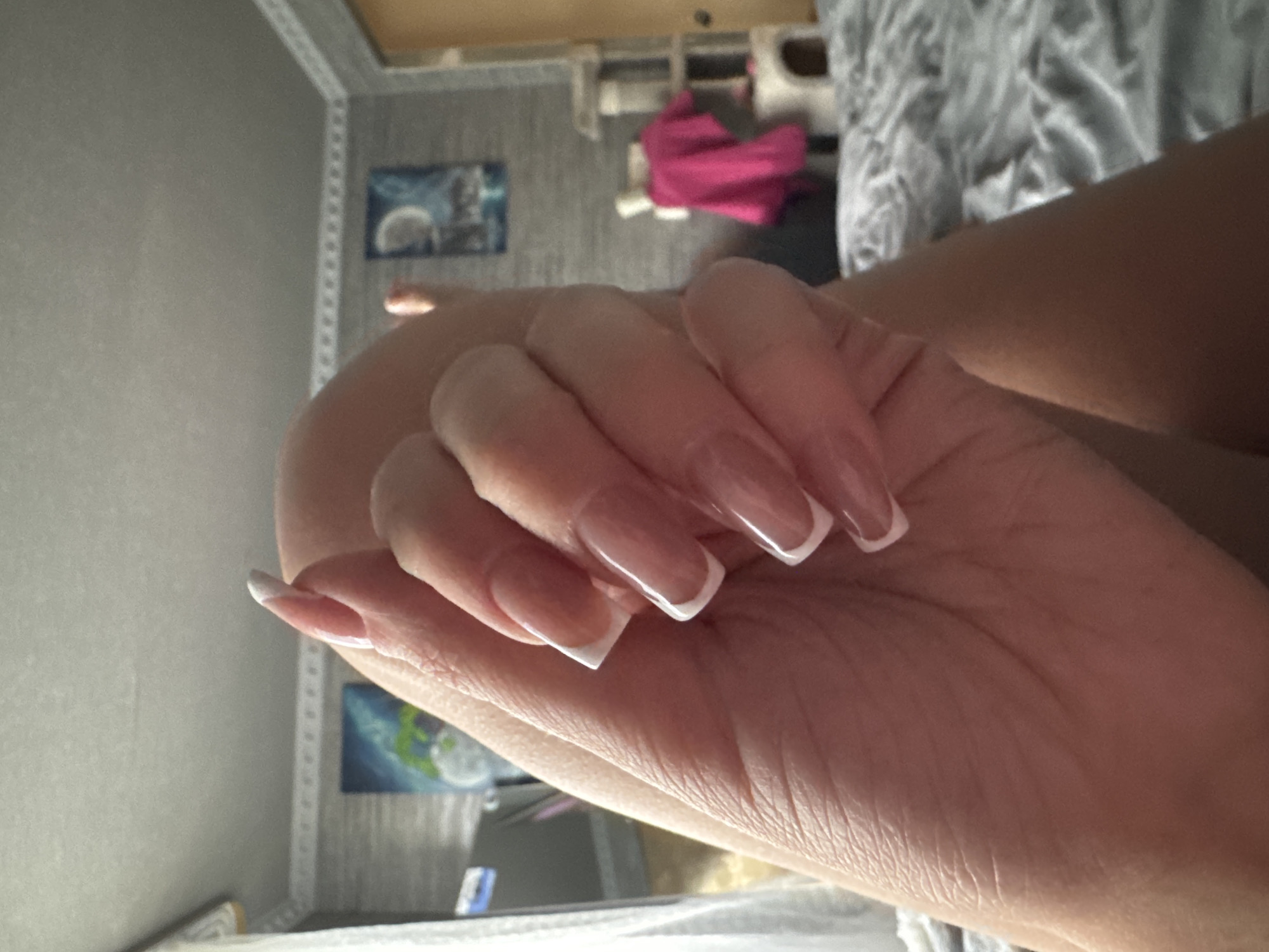 A close-up, over-the-shoulder shot shows a person's hand with French manicure acrylic nails resting on their leg, which is covered by a gray, textured blanket. The nails are long and curved, with a translucent pink base and a white tip. In the background, a room is visible, featuring a textured gray wall, a white decorative border along the ceiling, and two framed artworks depicting celestial scenes. A cat tree with pink fabric is also present, along with a door and other furnishings. The lighting suggests an indoor setting.