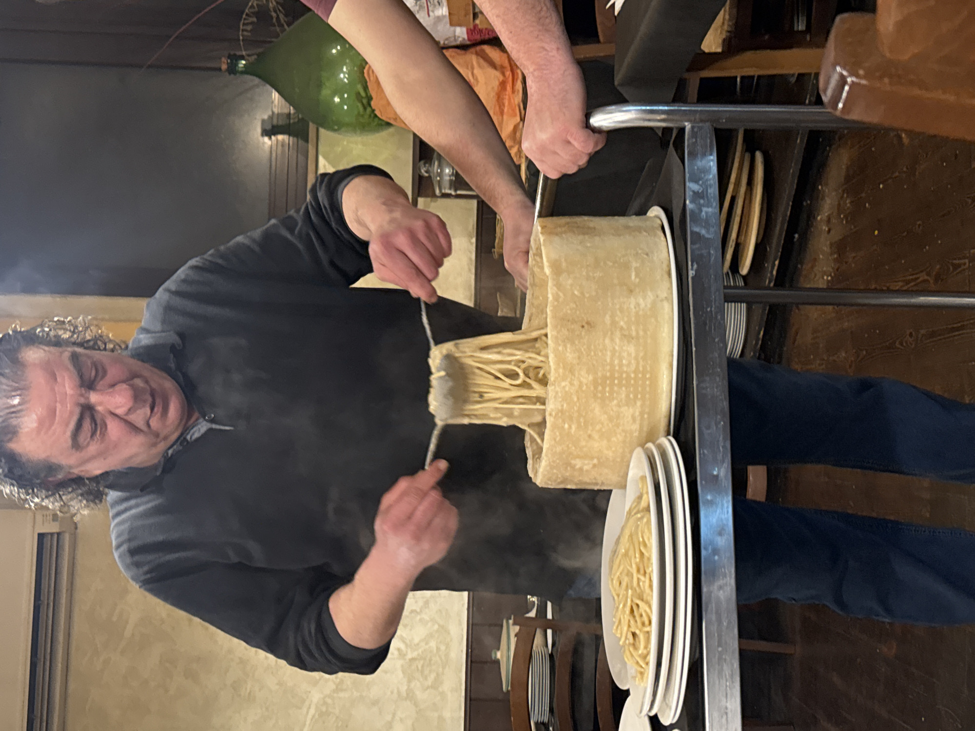 The image depicts an indoor scene within a restaurant, likely located in Rome, Italy. A middle-aged man with dark, curly hair, wearing a black polo shirt and blue jeans, is centrally positioned, actively serving pasta. He uses metal tongs to transfer steaming pasta from a large, hollowed-out wheel of cheese, which contains a substantial quantity of pasta coated in a creamy sauce. Visible steam rises from the cheese wheel and pasta.
To the man's left, a stack of white plates rests on a black-surfaced serving cart, with the top plate already containing a portion of the sauced pasta. The cheese wheel is also placed on this cart, which features a silver-colored metal frame. To the right, the forearm and hands of a second individual, wearing a reddish-brown long-sleeved shirt, are visible, resting on the metal frame of the serving cart. This individual appears to be observing the serving process.
The background on the left features a light-colored, textured wall with an air conditioning unit mounted high. A stack of white plates and part of a wooden chair are visible further left. On the right, a large, green glass demijohn bottle is prominent against a darker background, with reflections suggesting further depth. The floor is composed of dark wooden planks. The scene portrays a tableside pasta preparation and serving experience.