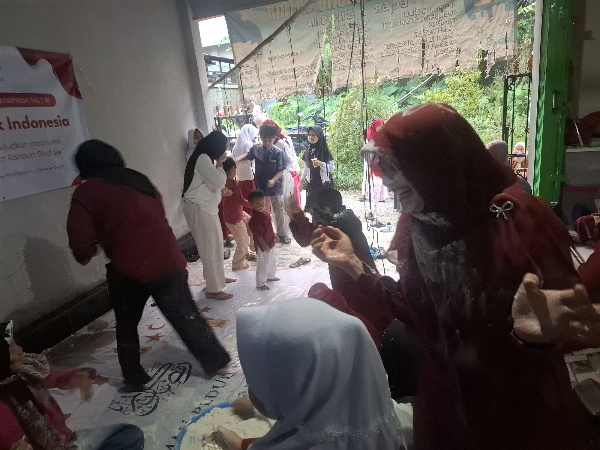 In an indoor-outdoor space in Babakan Madang, Indonesia, a group of people, including adults and children, are gathered for an event, possibly a celebration or a game. The floor is covered with a white plastic sheet decorated with Arabic calligraphy and crescent moon and star motifs. In the foreground, a person wearing a red head covering and dark clothing is covered in white powder. To their left, another person in a light blue head covering is crouched down, interacting with the powder on the floor. Several other individuals are visible in the background, some wearing white and red clothing. A banner with red and white elements is partially visible on the left, featuring text that appears to be in Indonesian. Behind the group, a cluttered outdoor area with greenery and what looks like a motorcycle suggests the location is a backyard or an open-air venue. The overall scene is dynamic, with people engaged in various activities, and the presence of powder suggests a playful or celebratory atmosphere.