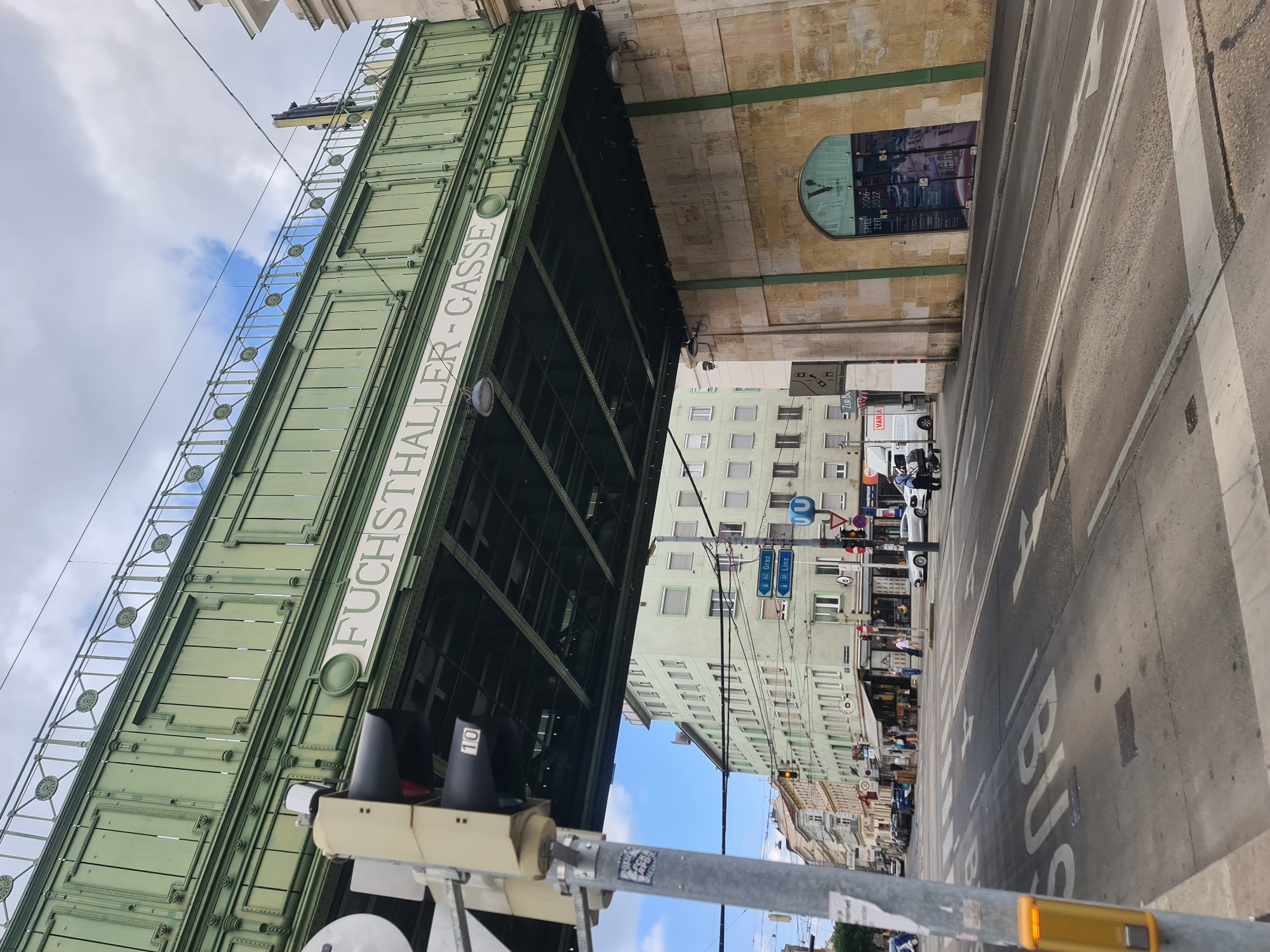 An eye-level shot captures a street scene in Vienna, Austria, under a green, elevated structure that displays the text "FUCHSTHALLER-CASSE" in white lettering. This structure appears to be part of the city's U-Bahn (subway) system, as indicated by a blue U-symbol sign visible on a pole to the right. The scene is dominated by the green metalwork of the overpass, supported by a substantial stone pillar on the right.
In the foreground, a street with multiple lanes and tram tracks runs horizontally. White lines and arrows mark the road surface, including a "BUS" lane marking. A traffic light with black lamps and a white number "10" is prominent on the left. A yellow pedestrian crossing button is mounted on a pole to the far left.
On the left side of the street, several cars are parked along the curb. Pedestrians can be seen walking on the sidewalk and entering shops. Buildings line the street, with a pale green multi-story building being the most noticeable. Signs for "GRAZ" and "LINZ" suggest road directions.
On the right, the stone pillar supporting the overpass is adorned with a large arched window displaying posters or advertisements. One poster has the text "SPIEL 2026 ZEIT 2027." A white vehicle, possibly a van, is parked on the right side of the street.
The sky above is partly cloudy with patches of blue. The overall impression is of a busy urban environment with functional public infrastructure.