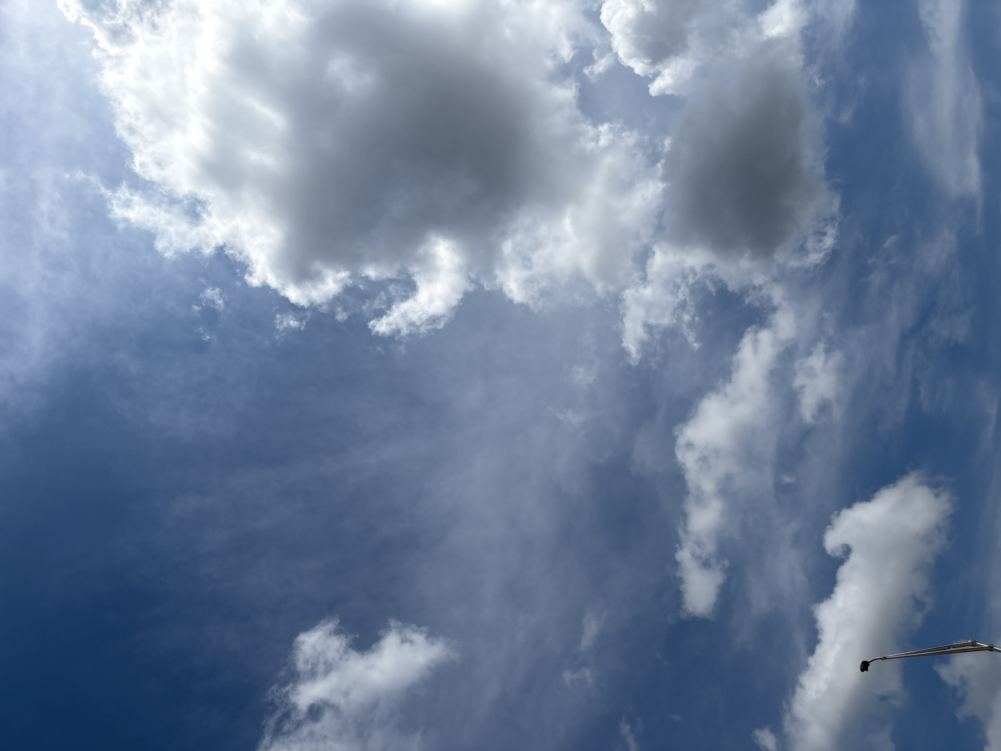 A wide-angle, vertically oriented, medium shot captures a bright, cloudy blue sky from a low-angle perspective. Dominating the right side of the frame, a large, dense cumulus cloud formation features stark white highlights and deep gray shadows, indicating significant depth. Smaller, wispy clouds, appearing as white streaks, are scattered across the upper left and lower sections of the image. The lower left corner shows the top of a metal lamppost, its angled shaft extending upwards towards the sky. The scene is likely outdoors in Duette, United States, given the clear, expansive sky and the presence of a lamppost, consistent with an outdoor setting.