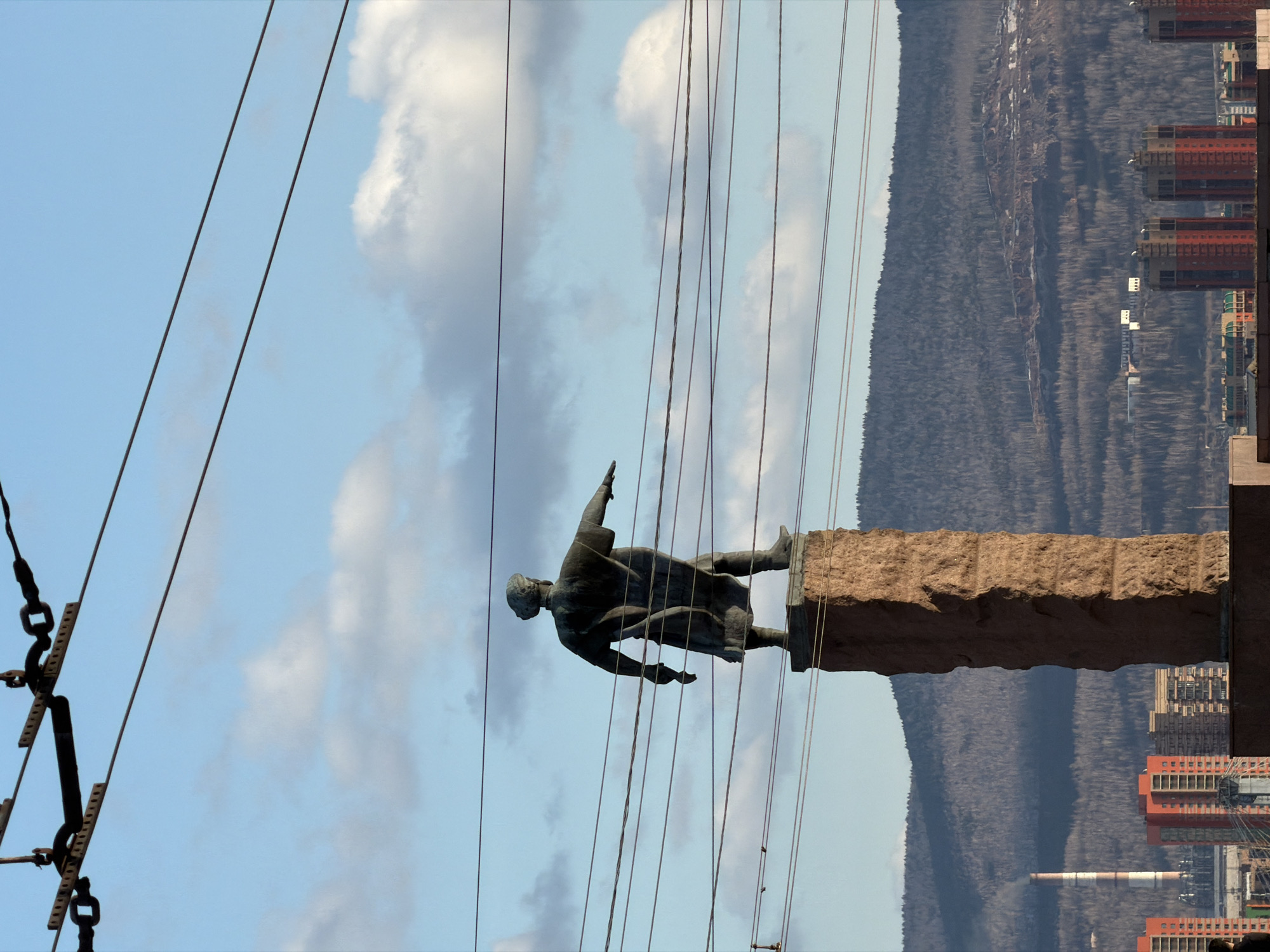 The image shows a sideways view of the monument to Nikolay Ostrovsky in Krasnoyarsk, Russia. The bronze statue of Ostrovsky stands atop a rough-hewn stone pedestal. Power lines are visible against a backdrop of clouds and sky. A wooded hillside is visible behind the statue, with a cityscape featuring red brick buildings and a smokestack visible in the distance. A utility pole structure can be seen to the left.