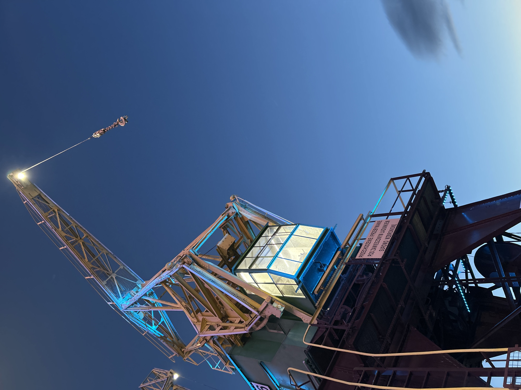 The image shows a close-up of a yellow gantry crane at dusk, with a clear blue sky in the background. The crane is illuminated with blue and yellow lights. A sign in Polish is visible on the lower structure of the crane. This is likely the historic DOK 7 crane located in the Wały Chrobrego area of Szczecin, Poland, which is part of the National Maritime Museum. There are no people visible in the image.