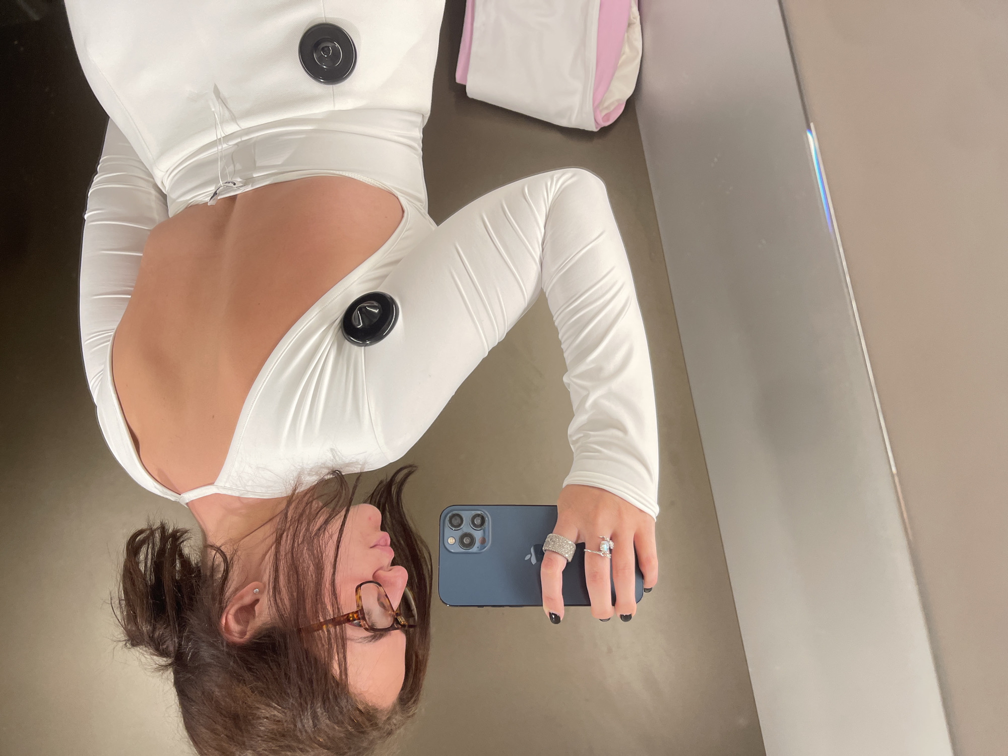 The image shows a person taking a selfie in a location with beige and grey walls, possibly a dressing room or bathroom. The person is wearing a white, long-sleeved garment with large, black button-like decorations. The garment is cut out in the back. The person is holding a blue smartphone, adorned with multiple rings on the fingers holding the phone. The person has brown hair and is wearing glasses. A pink and white object is visible in the background. The location is not identifiable from the image. Therefore, it cannot be determined whether it is in the city of Sovkhoza Imeni Lenina, Russia.