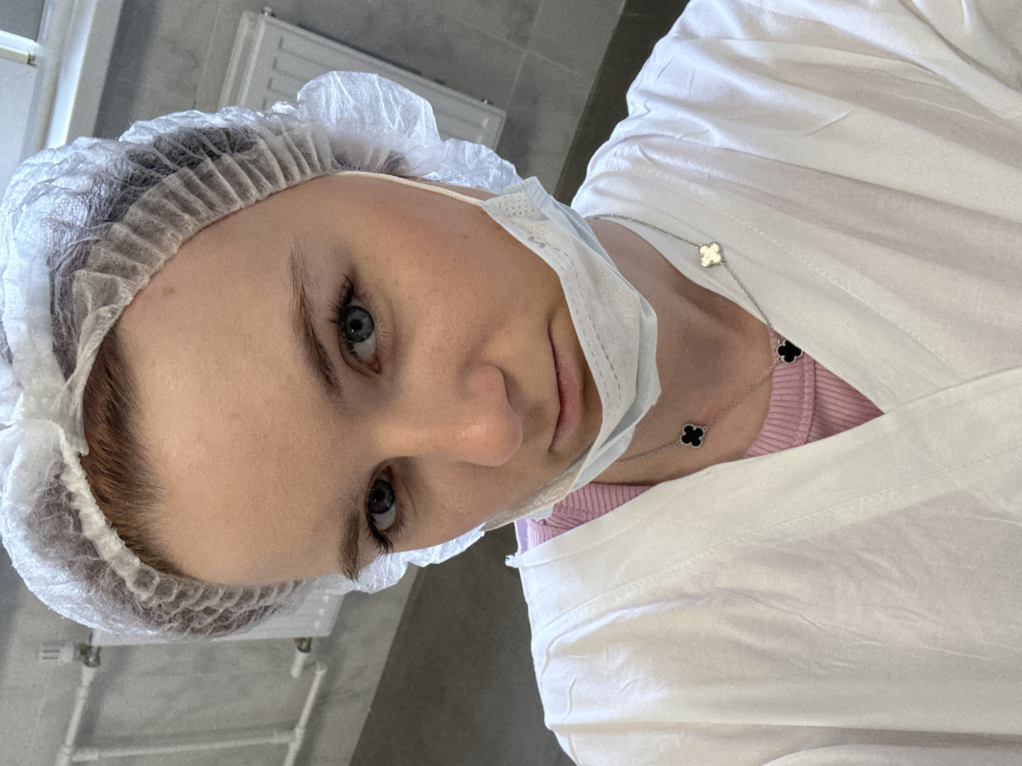 A young woman is seen indoors, likely in a medical setting, as she wears a white bouffant cap and a surgical mask pulled down below her chin. She is also wearing a white lab coat over a pink, possibly ribbed, undershirt. Around her neck is a delicate necklace with clover-shaped pendants in black and white.
The background features neutral colors, including white walls and gray flooring. A radiator is visible in the upper left corner. The woman's face is the primary focus of the image, showing light skin, blue eyes, and dark eyebrows.
