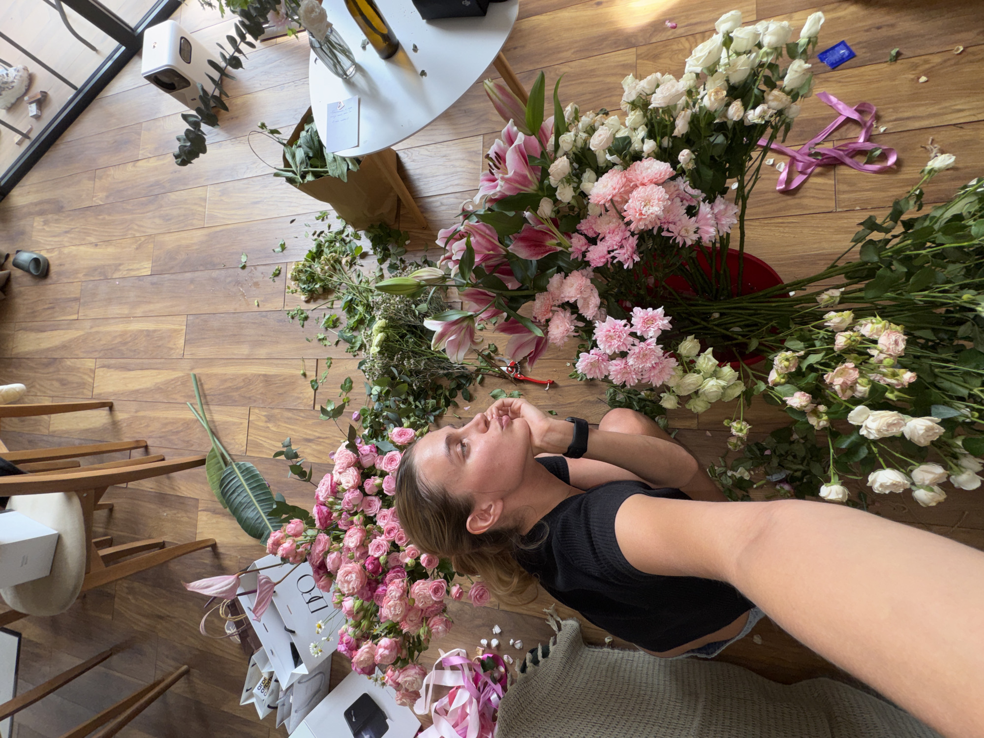 A high-angle, slightly tilted shot captures a woman lying on a light brown wooden floor, surrounded by an abundance of cut flowers and foliage. The indoor setting features natural light, likely from a large window or glass door with a black frame visible in the background.
The woman, with blonde hair and fair skin, is dressed in a black top and dark shorts. She lies with her head propped on her left arm, looking upwards with a calm, contemplative expression. A black smartwatch is visible on her wrist.
The floor is covered with numerous flower varieties, predominantly in shades of pink and white. These include pink and white roses, pink lilies, and pink chrysanthemums, along with various green leaves and stems. A red pair of floral shears is placed among the flowers, and scattered pink ribbons add to the scene.
Other objects in the environment include a small white round table with a bottle and a vase, a brown paper bag, and several white shopping bags. One shopping bag clearly displays the text "D.O.". A white gift box, a black object (possibly a phone), and a grey textured blanket are also visible. In the background, a white, elongated device, possibly an air purifier, is mounted on a wall or window frame, and a dark object (possibly a speaker) is on the floor. A distinct blue wrapper, resembling a condom wrapper, is visible on the wooden floor near a cluster of white flowers and pink ribbon.
The scene conveys a relaxed, perhaps artistic, daytime activity centered around flower preparation or enjoyment.