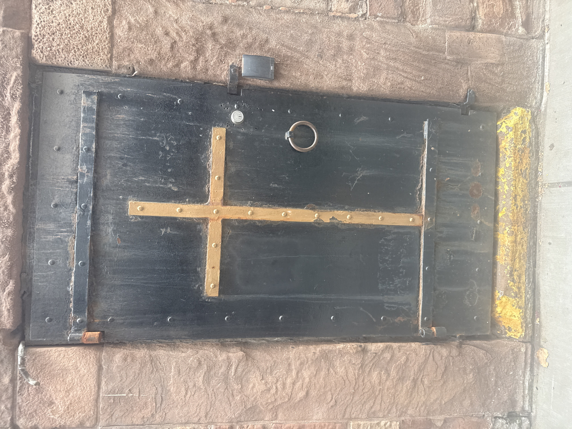 The image shows a closed, heavy, black metal door set within a rough, reddish-brown stone wall. A golden cross is affixed to the center of the door, composed of two rectangular planks joined together, with gold-colored rivets securing them. The door has a metal ring handle and a keyhole lock. To the right of the door, mounted on the stone wall, is a small black electronic access control device. The bottom of the door shows signs of wear, with chipped yellow paint along the threshold. The ground in front of the door appears to be a sidewalk made of concrete, with some fallen leaves. The overall impression is one of a sturdy, possibly historic entrance. The location is the City of New York, United States of America, specifically at the entrance to the crypt of Trinity Church.