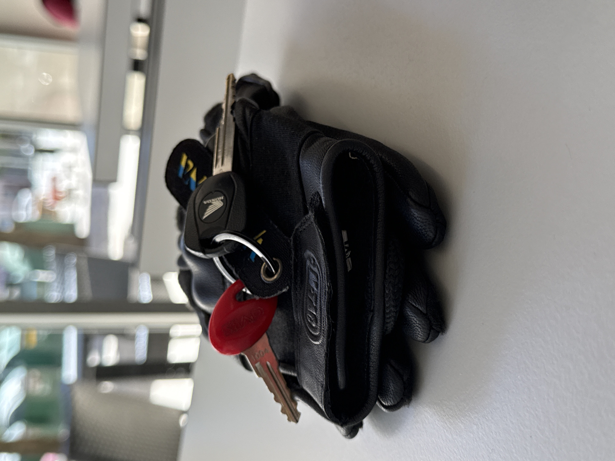 A pair of black leather motorcycle gloves, with the brand "ALPINESTARS" embossed on the palm area, are resting on a light gray, textured surface. Three keys are attached to a keyring on top of the gloves. One key has a red plastic fob, and another has a black fob with a yellow and blue emblem, and a Honda logo. The background is blurred, showing an indoor setting with windows and furniture, suggesting a cafe or public area. No people are visible in the image. The scene is devoid of any specific visual cues that would pinpoint its location to Pamplona, Spain.