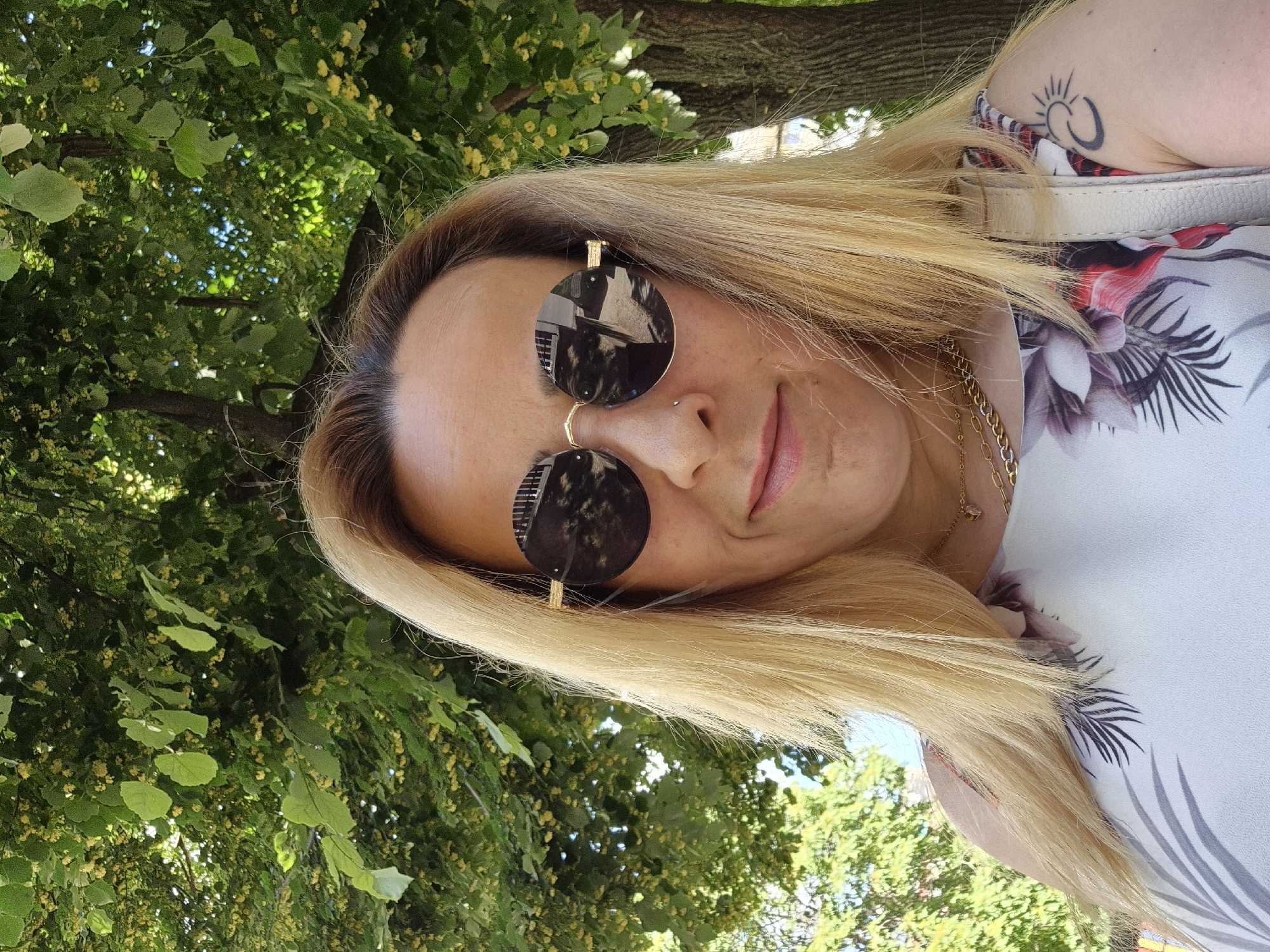 A medium shot captures a blonde woman with fair skin wearing round, dark sunglasses and a floral-patterned dress. She has a nose ring and is smiling slightly, with her mouth slightly ajar. A gold chain necklace is visible around her neck. A tattoo of a sun and cloud is on her left shoulder. She is outdoors, with a large tree and green foliage in the background. The lighting suggests it is daytime, possibly a sunny day. The environment appears to be a park or a tree-lined street.