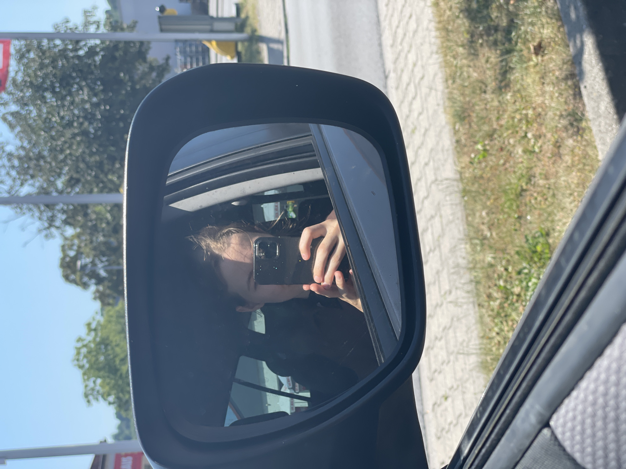 A person is taking a selfie in the side-view mirror of a car. The person's face is partially visible, as they are holding a black smartphone in their right hand. Their left hand is also visible, supporting the phone. The person appears to have light-colored hair and is wearing a dark jacket or shirt. The reflection shows the interior of the car, including the dashboard and a window. Outside the car, the scene includes a tree with green foliage against a clear blue sky on the left. On the right side of the mirror's reflection, a paved area, possibly a sidewalk or road, is visible next to a patch of dry grass. The overall lighting suggests it is daytime, likely sunny. The location context is Nidda, Germany, which is a town in the state of Hesse. The image has a casual, everyday feel, possibly a moment captured while driving or stopped in the car. No discernible text is visible in the image.