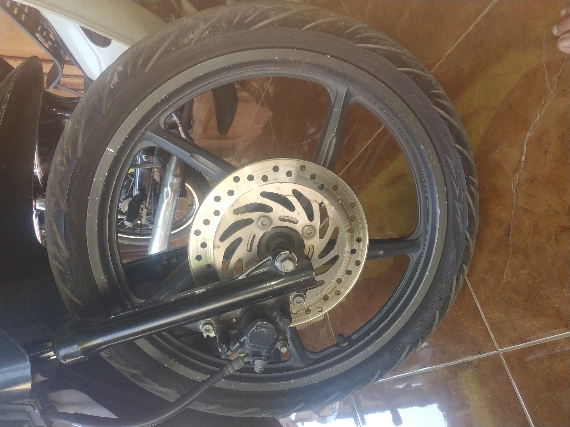 The image shows a close-up, low-angle shot of a motorcycle's front wheel and braking system, placed on a tiled floor. The motorcycle is black and appears to be stationary, possibly parked indoors or in a covered area, as indicated by the tiled floor which often features in residential or commercial spaces in Jember, Indonesia.
The primary subject is the front wheel, featuring a black rim with a patterned tire. A silver disc brake is visible in the center of the wheel, with a black brake caliper mounted to the left of the disc. The motorcycle's black front suspension fork is also prominently displayed, extending upwards out of frame. The background is blurred, but suggests the presence of other parts of the motorcycle, possibly another wheel, and some light-colored paneling, as well as wooden elements. The lighting suggests an indoor environment, possibly with natural light filtering in, creating reflections on the polished tiled floor. The image has a slightly grainy texture, suggesting it was taken with a mobile device or a lower-resolution camera. The overall scene depicts a detailed view of the mechanical components of a motorcycle.